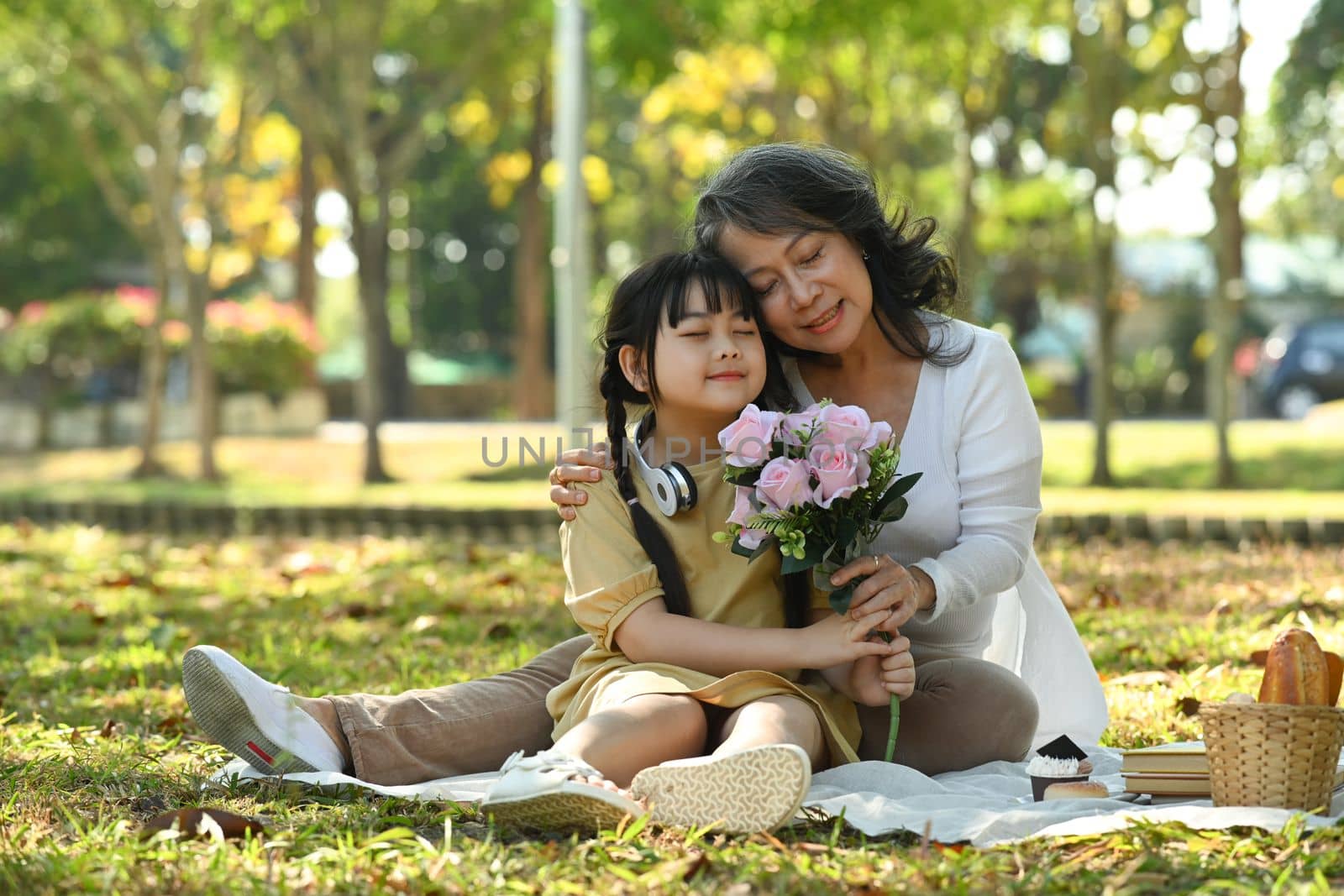 Caring senior woman hugging cute little grandchild while sitting on green grass in the park. Family, generation and love concept.