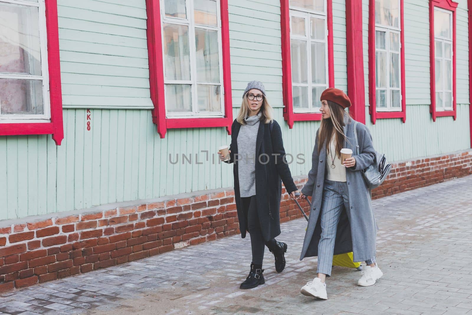 Two cheerful tourist women smiling and walking with suitcases on city street in autumn or spring time - travel and vacation