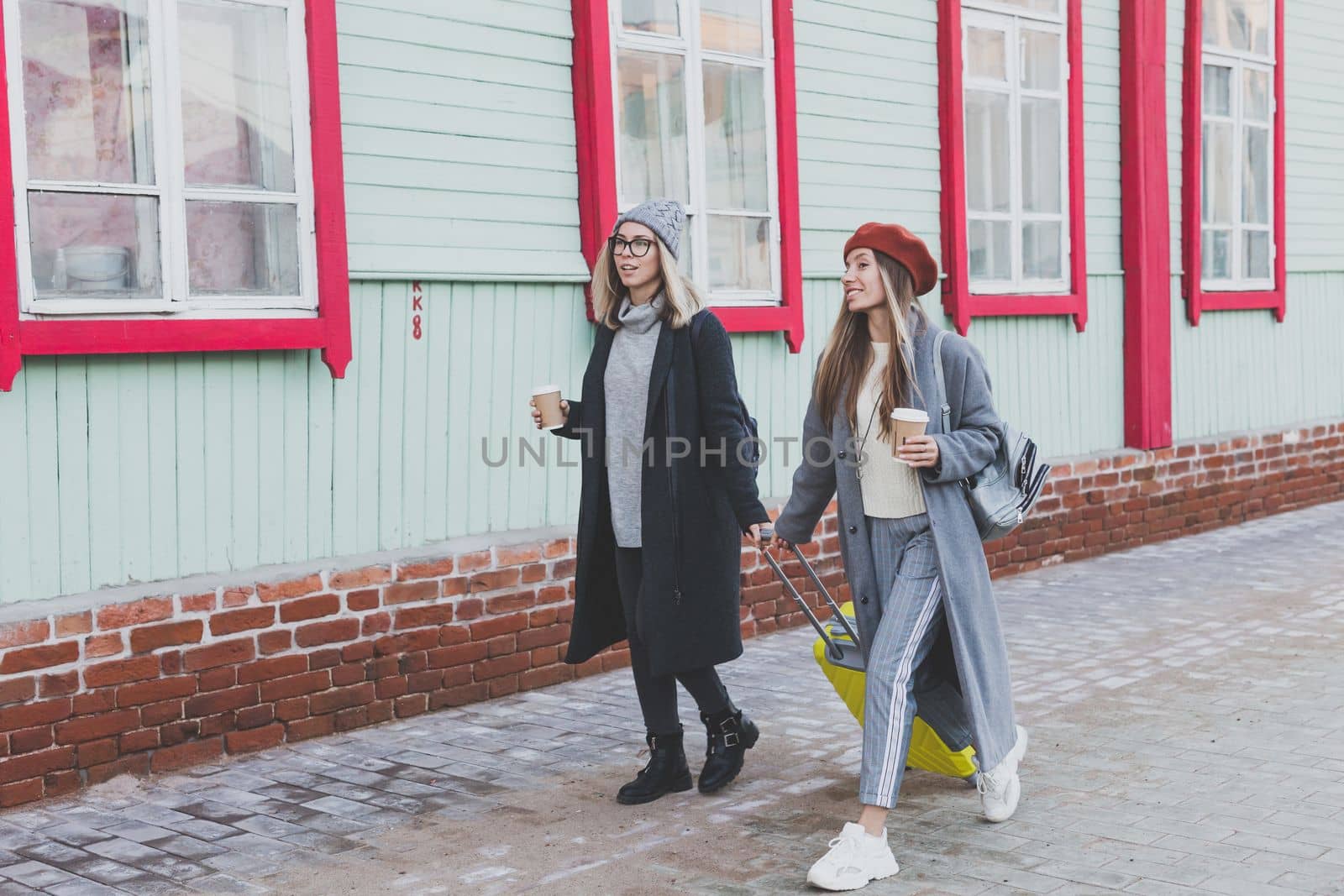 Two cheerful tourist women smiling and walking with suitcases on city street in autumn or spring time - travel and vacation concept by Satura86