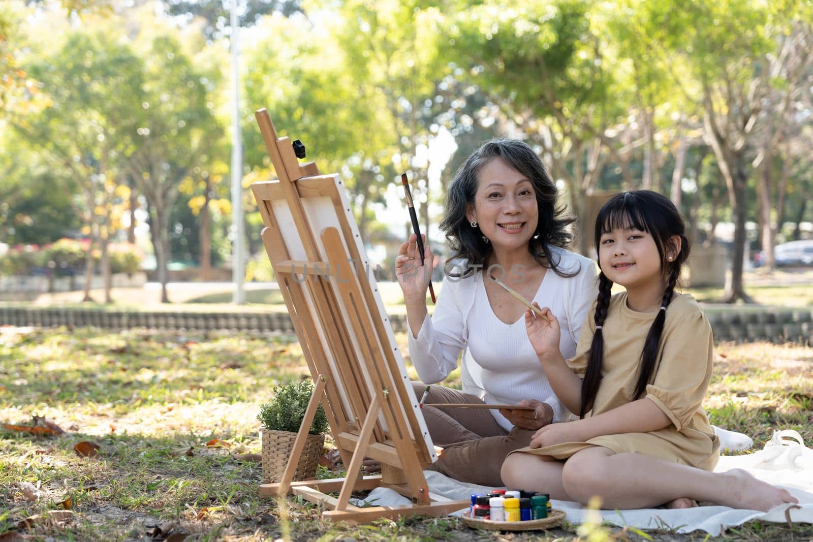 Happy and lovely Asian grandmother teaching her granddaughter to paint watercolors on canvas, having a great time together in the beautiful park.