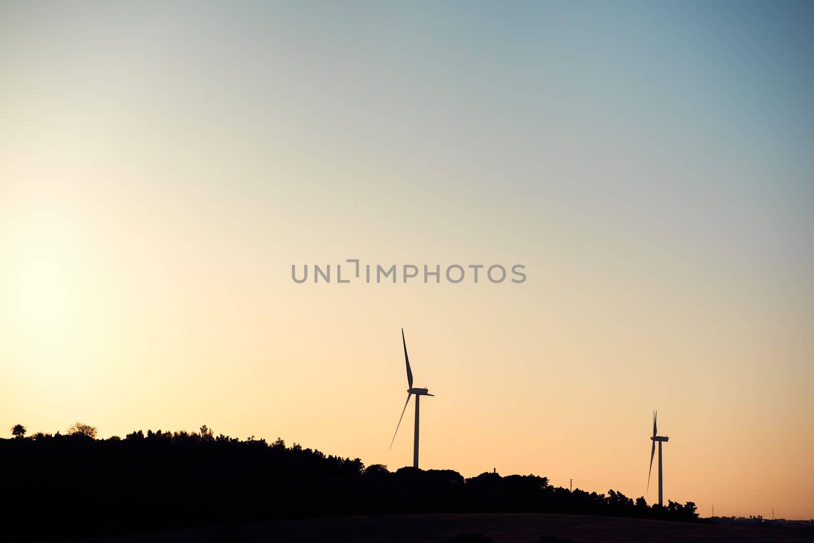 Silhouette of two windmills producing clean energy in a wind farm at sunset. They are in a rural environment surrounded by crops and trees, the sky is clean and clear. Copy space