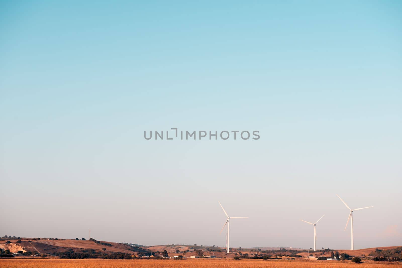 three windmills produce clean energy in a wind farm. They are in a rural environment surrounded by crops and small hills, the sky is clean and clear. Copy space