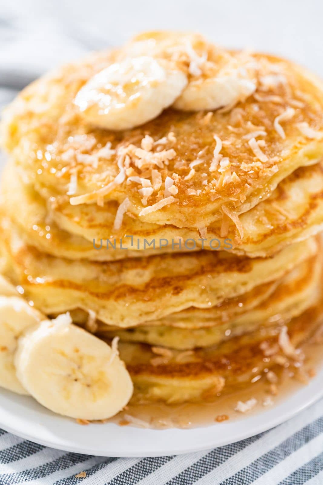 Stack of freshly baked coconut banana pancakes garnished with sliced bananas, toasted coconut, and maple syrup.