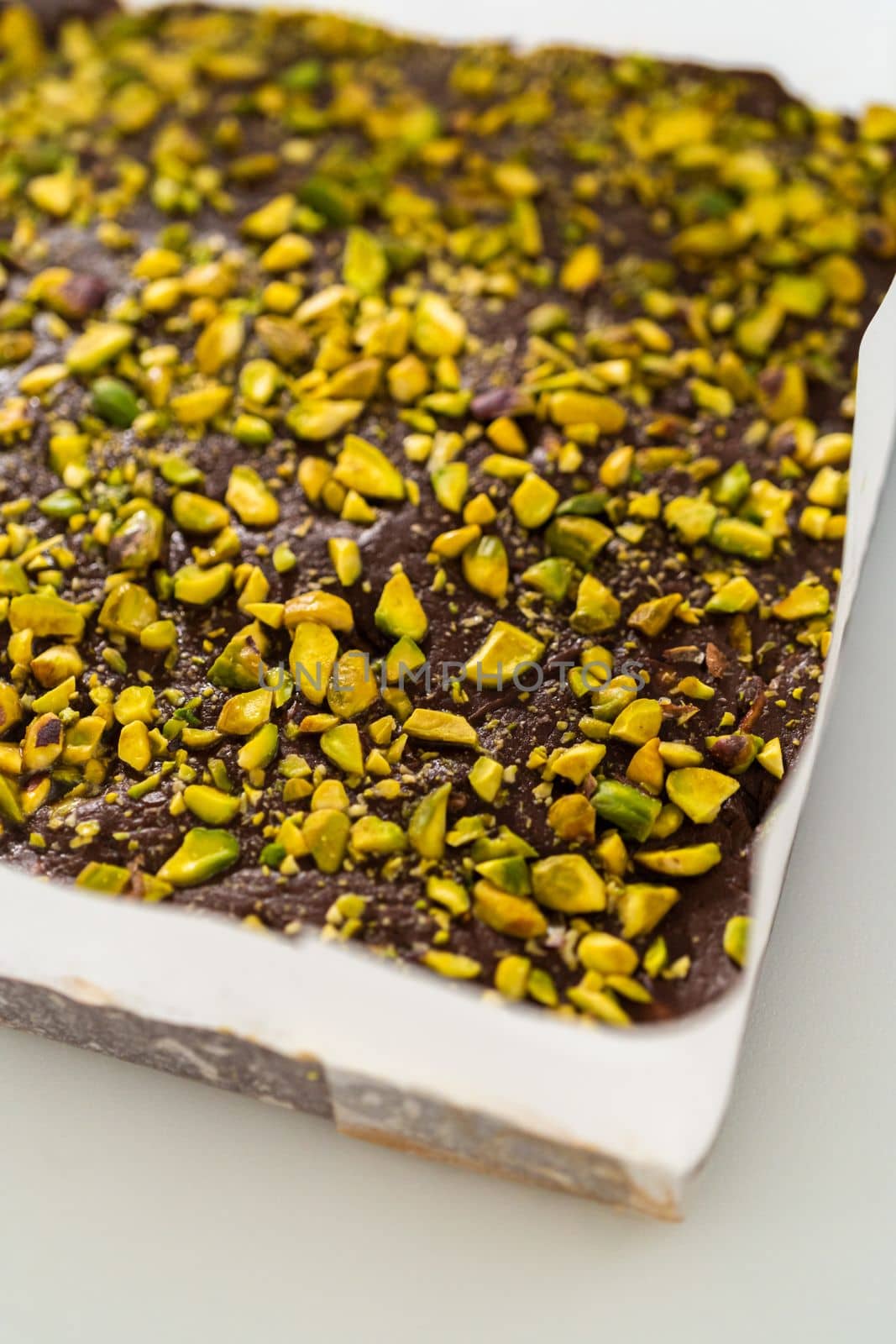 Removing chocolate pistachio fudge from a square cheesecake pan lined with parchment.