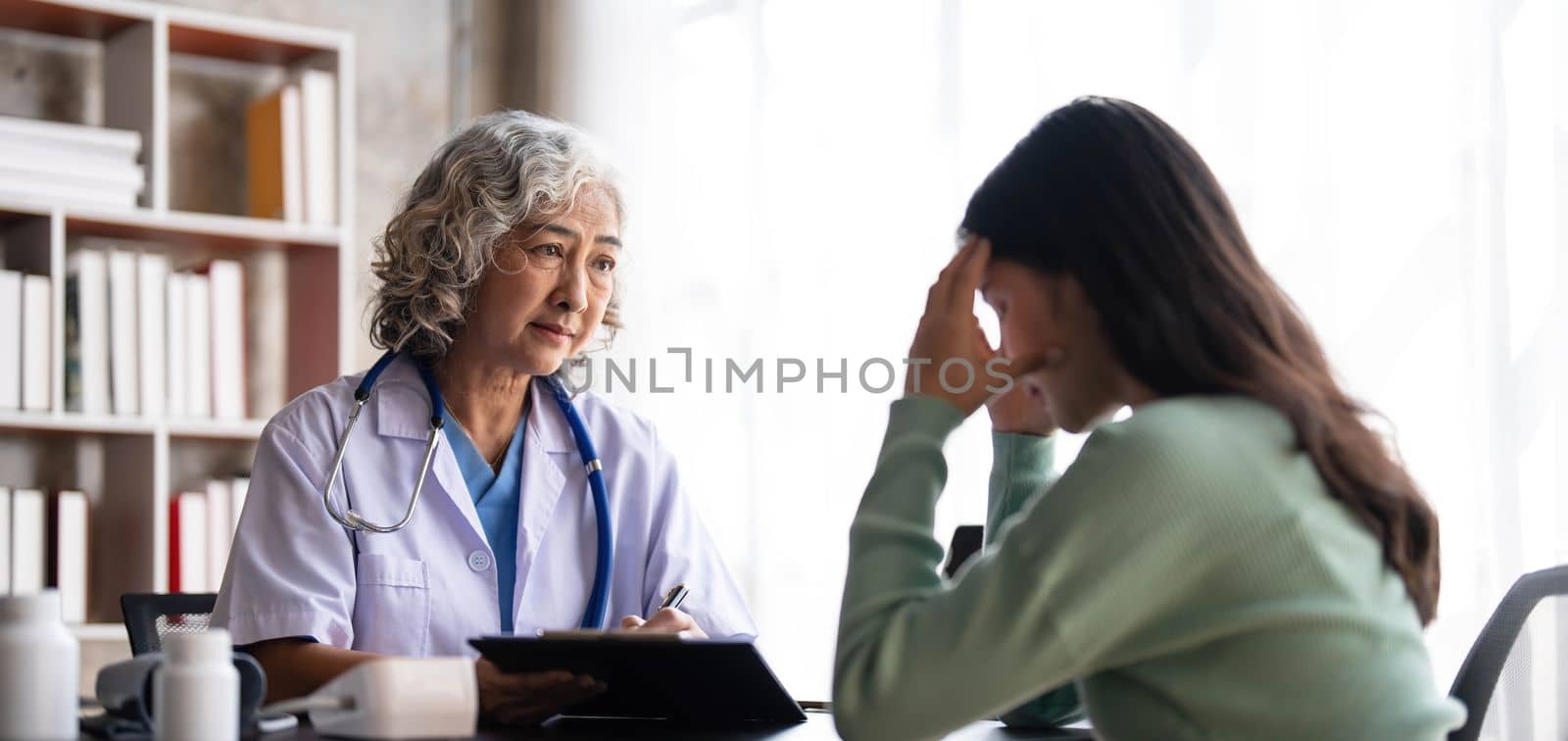 Woman senior doctor is Reading Medical History of Female Patient and Speaking with Her During Consultation in a Health Clinic. Physician in Lab Coat Sitting Behind a Laptop in Hospital Office...
