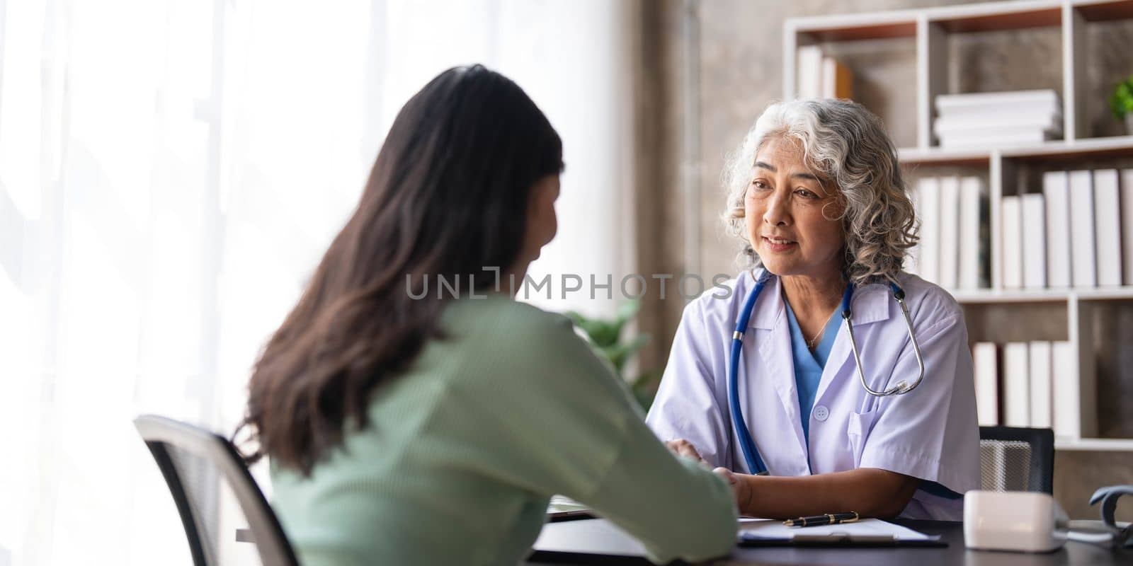 Woman senior doctor is Reading Medical History of Female Patient and Speaking with Her During Consultation in a Health Clinic. Physician in Lab Coat Sitting Behind a Laptop in Hospital Office...