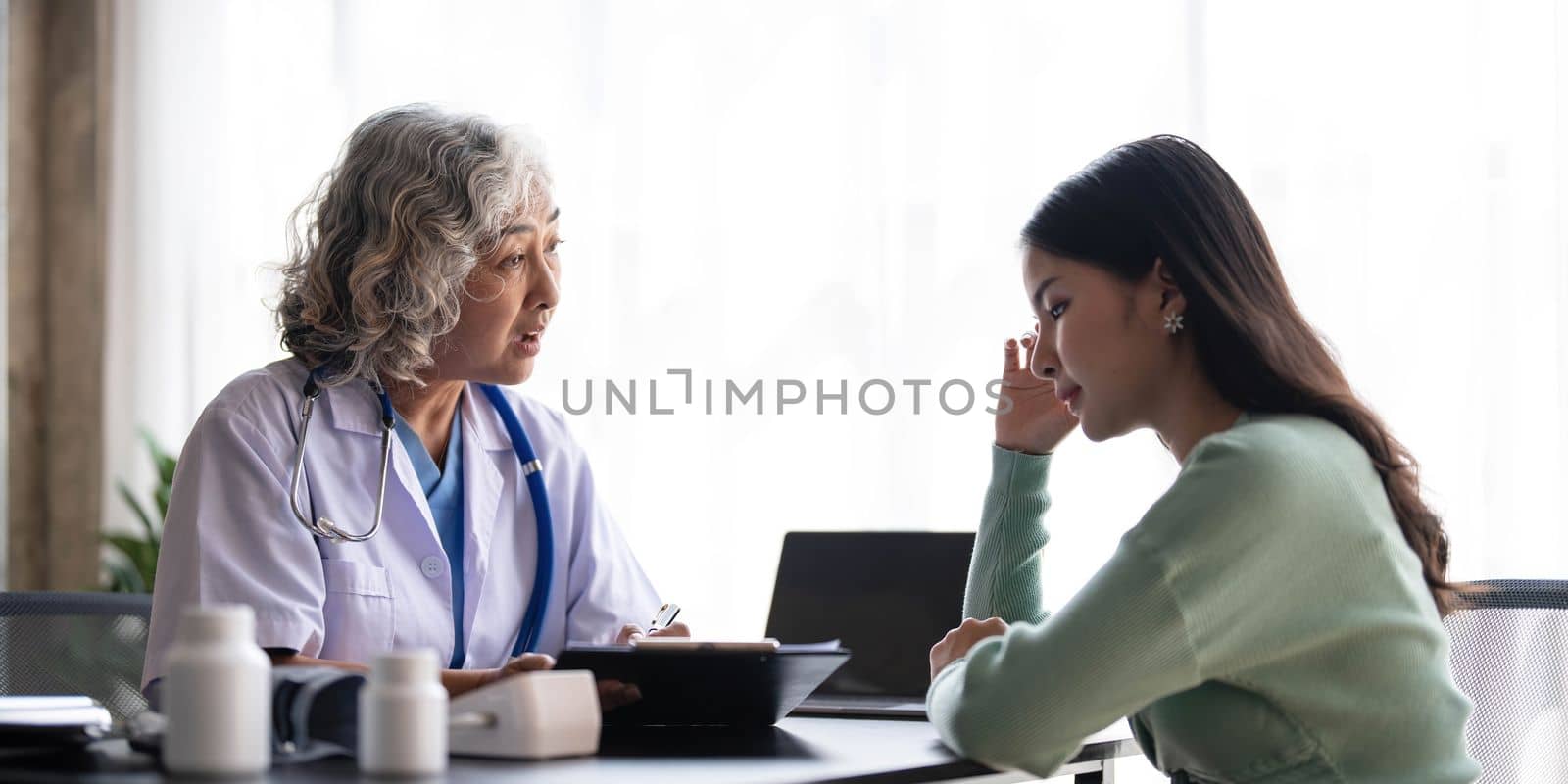 Woman senior doctor is Reading Medical History of Female Patient and Speaking with Her During Consultation in a Health Clinic. Physician in Lab Coat Sitting Behind a Laptop in Hospital Office. by wichayada