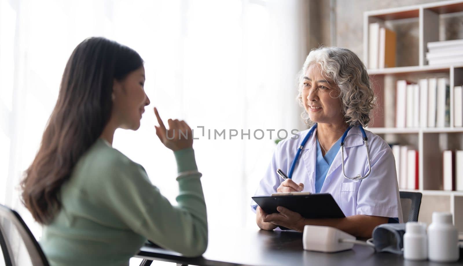 Woman senior doctor is Reading Medical History of Female Patient and Speaking with Her During Consultation in a Health Clinic. Physician in Lab Coat Sitting Behind a Laptop in Hospital Office. by wichayada