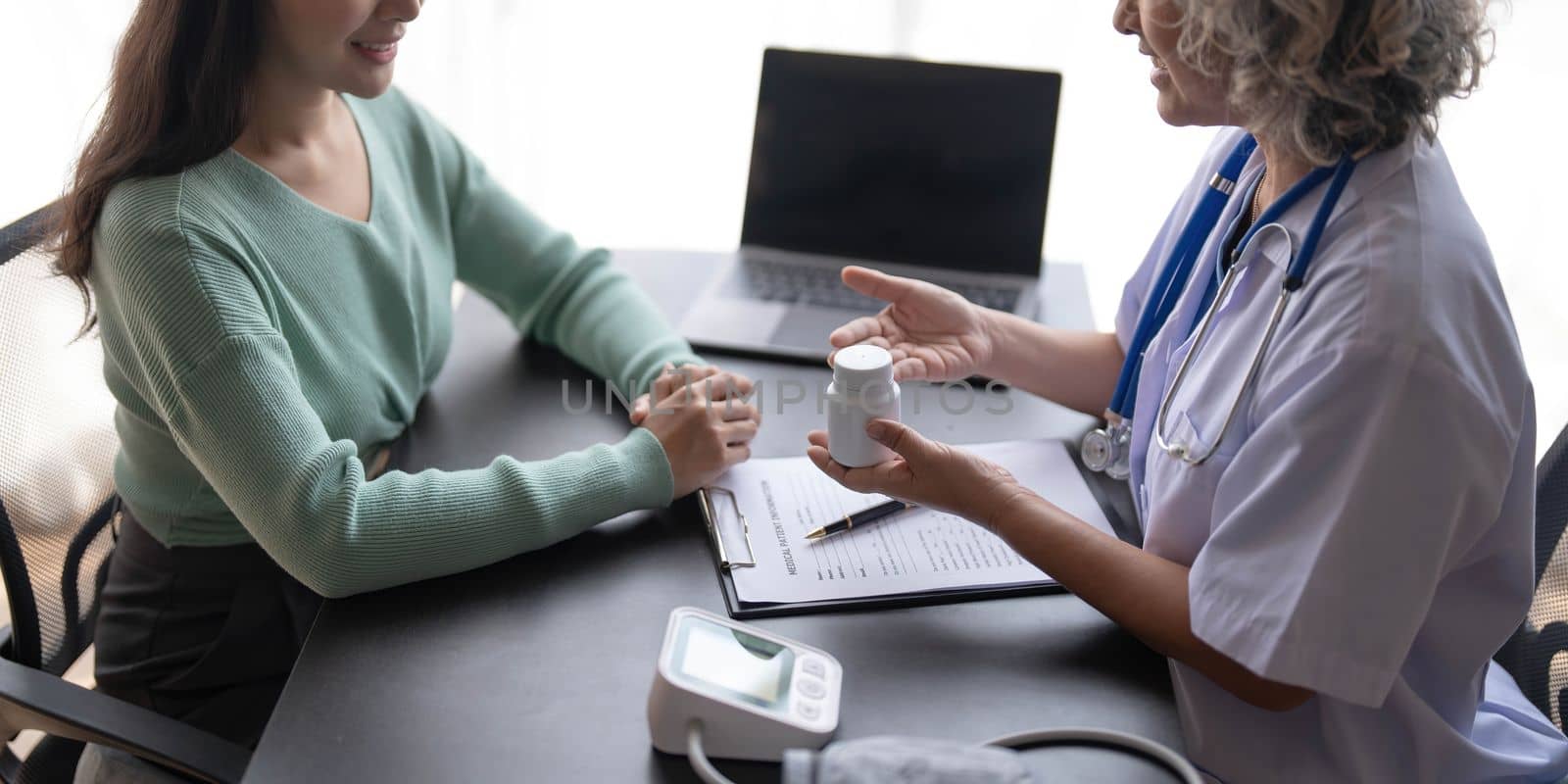 Woman senior doctor is Reading Medical History of Female Patient and Speaking with Her During Consultation in a Health Clinic. Physician in Lab Coat Sitting Behind a Laptop in Hospital Office. by wichayada