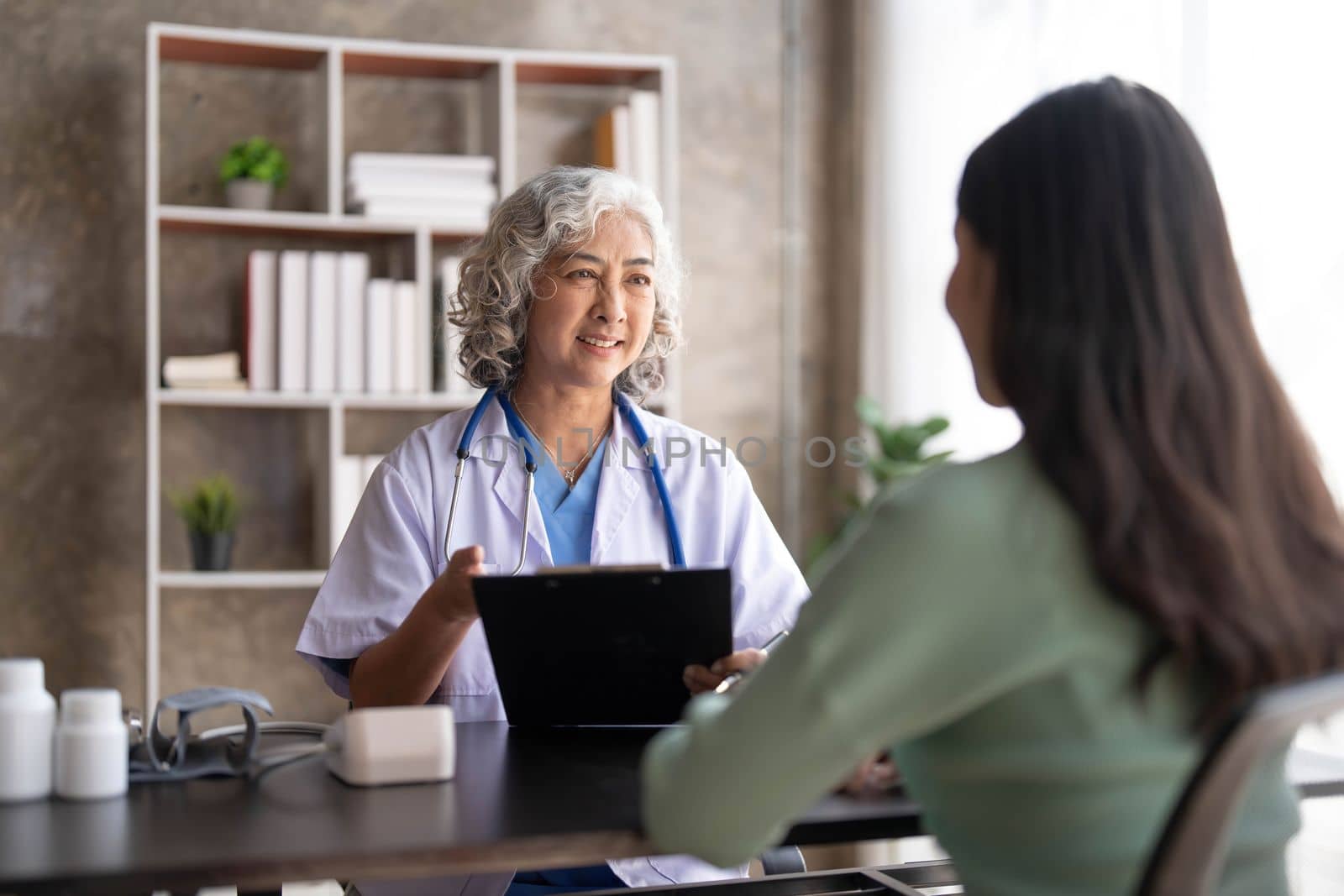 Woman senior doctor is Reading Medical History of Female Patient and Speaking with Her During Consultation in a Health Clinic. Physician in Lab Coat Sitting Behind a Laptop in Hospital Office. by wichayada