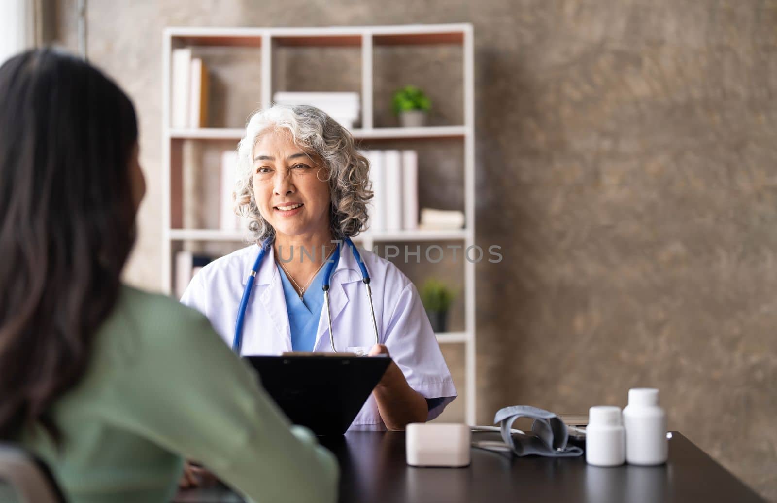 Woman senior doctor is Reading Medical History of Female Patient and Speaking with Her During Consultation in a Health Clinic. Physician in Lab Coat Sitting Behind a Laptop in Hospital Office. by wichayada