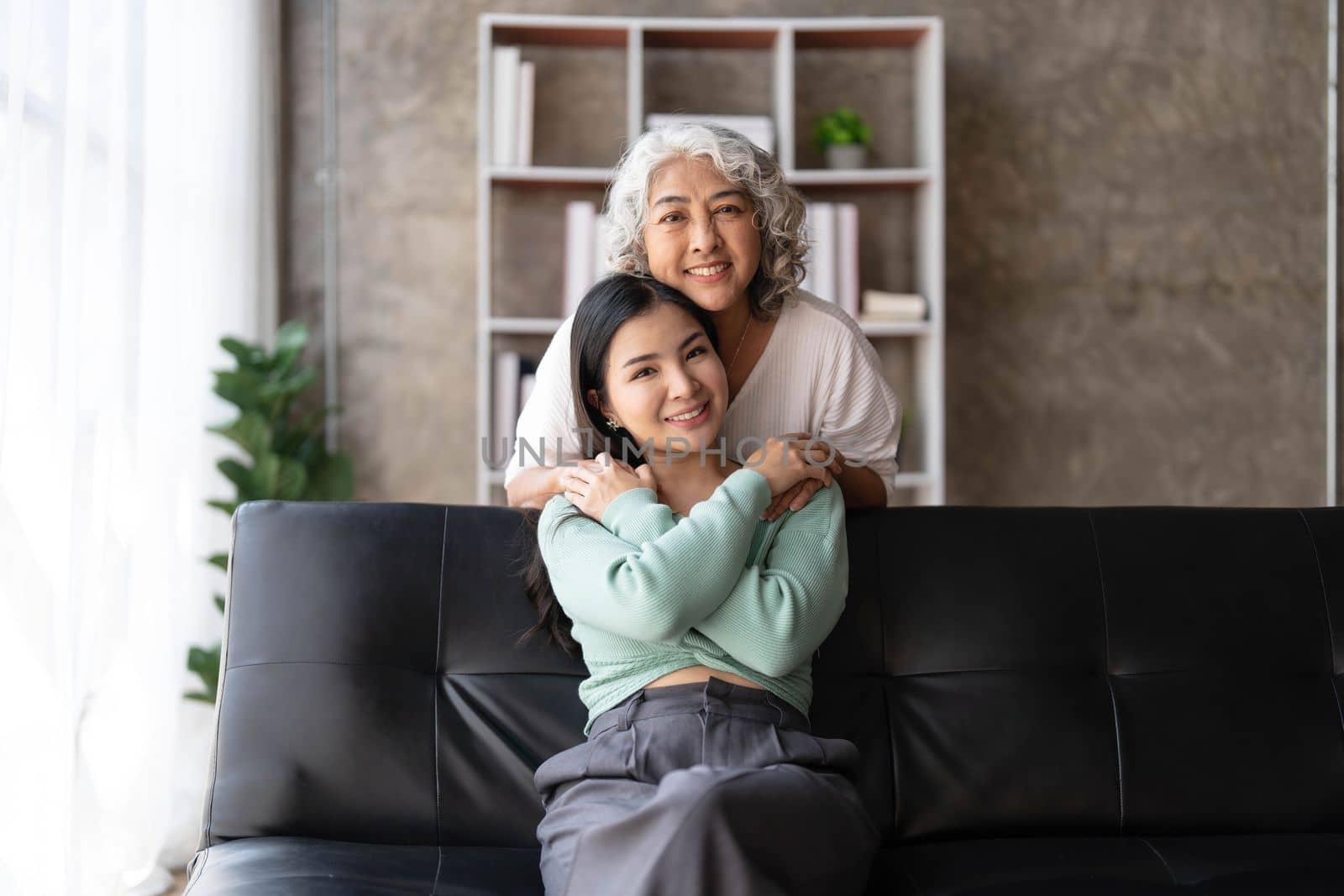 Loving adult daughter hugging older mother, standing behind couch at home, family enjoying tender moment together, young woman and mature mum or grandmother looking at each other, two generations..