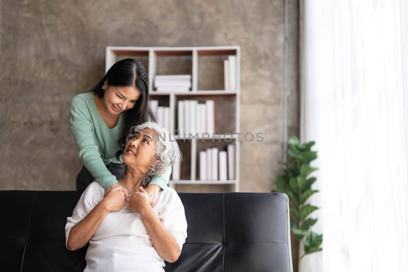 Loving adult daughter hugging older mother, standing behind couch at home, family enjoying tender moment together, young woman and mature mum or grandmother looking at each other, two generations..