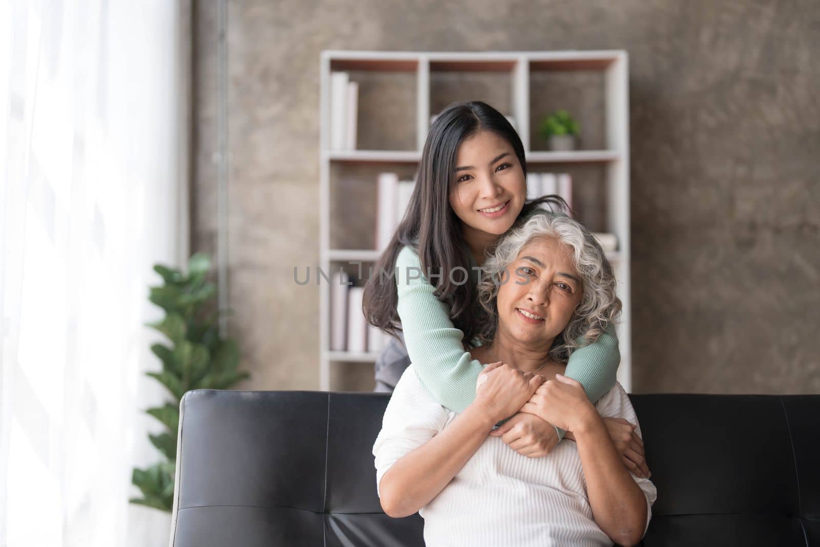 Loving adult daughter hugging older mother, standing behind couch at home, family enjoying tender moment together, young woman and mature mum or grandmother looking at each other, two generations by wichayada