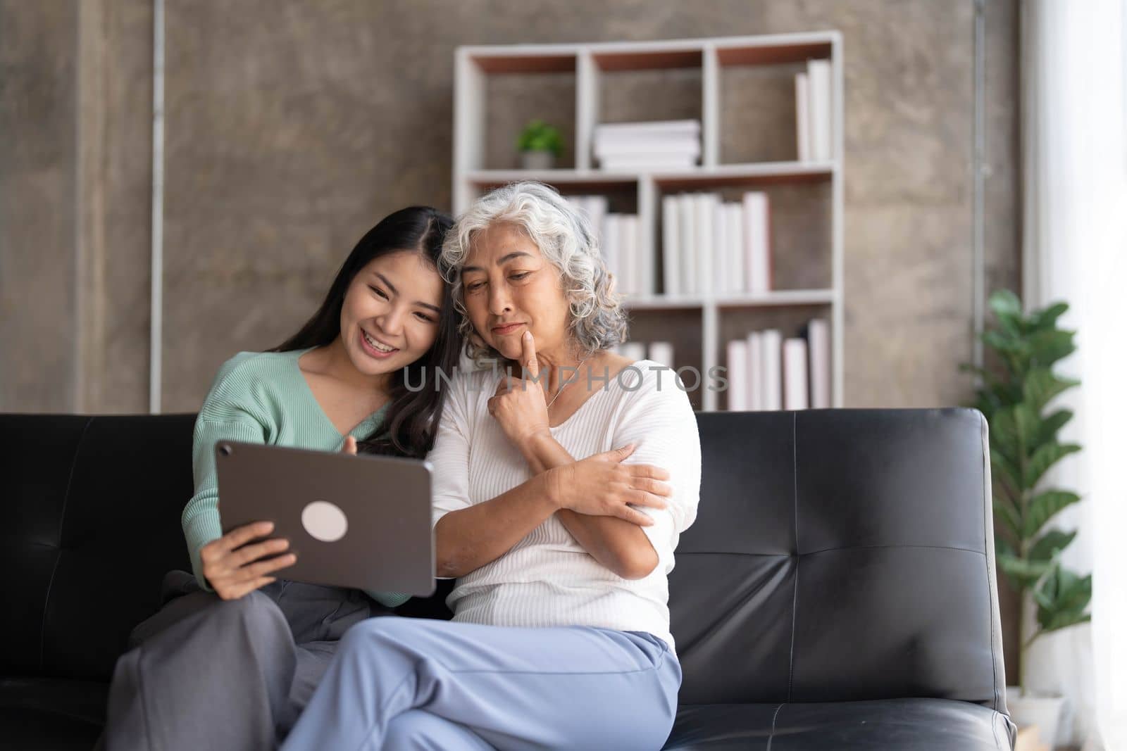 Mature mother grown up daughter sit on couch in living room hold on lap pc looking at screen watching movie film, using device make video call, shopping via e-commerce website, girl teach mom concept by wichayada