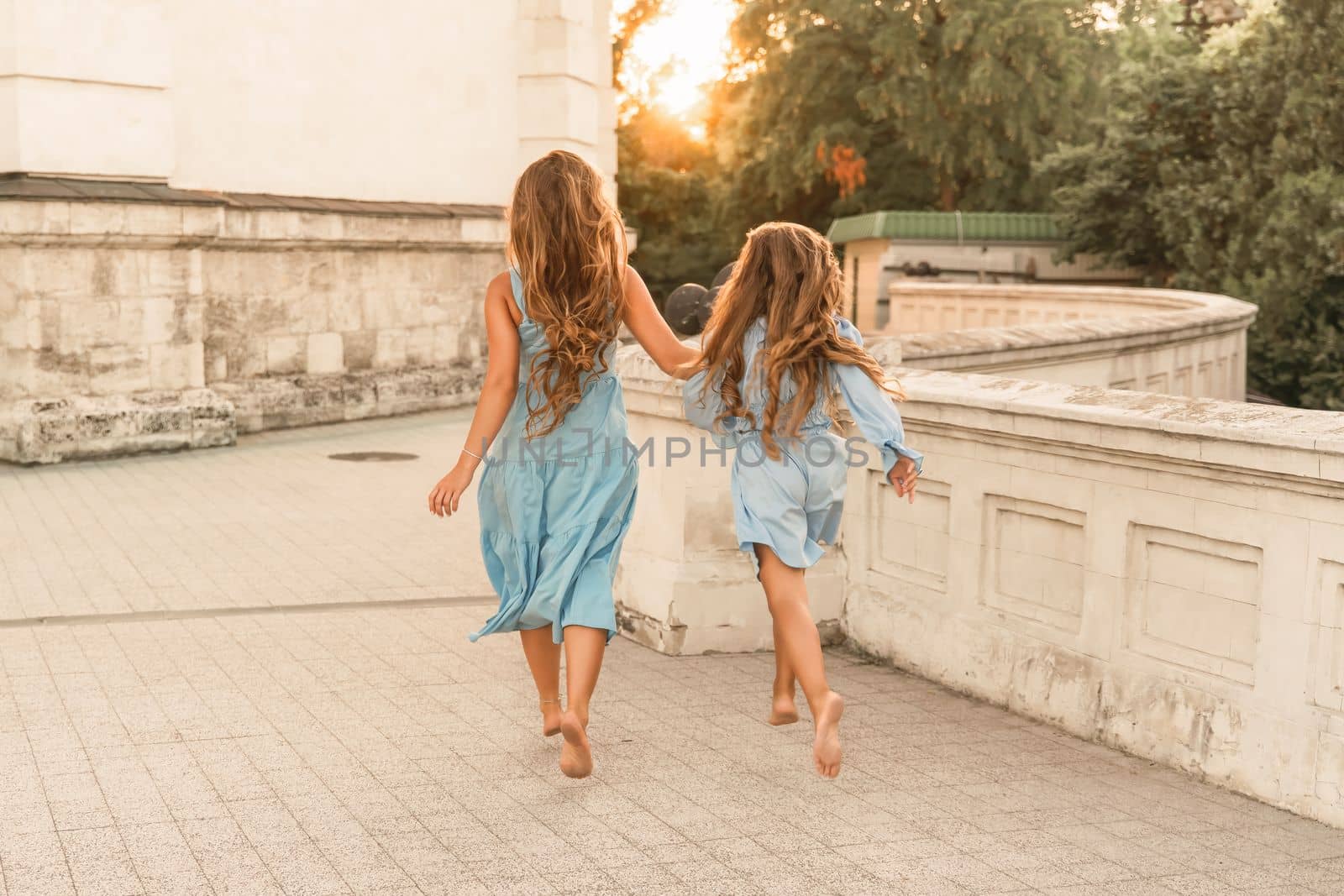 Daughter mother run holding hands. In blue dresses with flowing long hair against the backdrop of a sunset and a white building