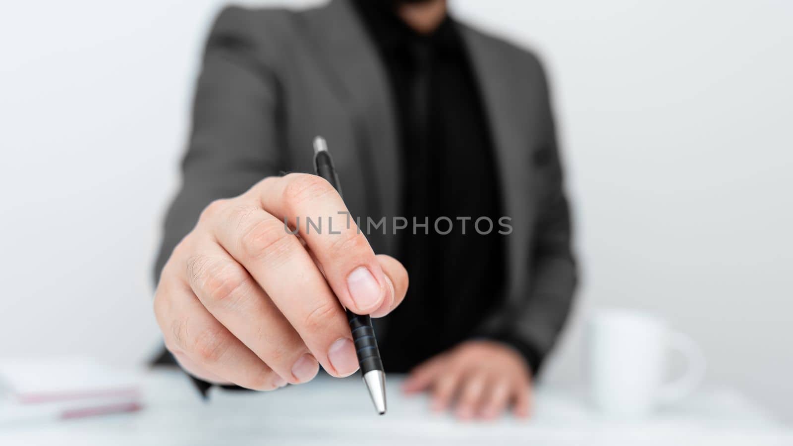 Male model in Gray suit sitting at white table And Pointing With Pen On Important Message. Gentleman Showing Critical Announcement. Coffee cup on deck. by nialowwa
