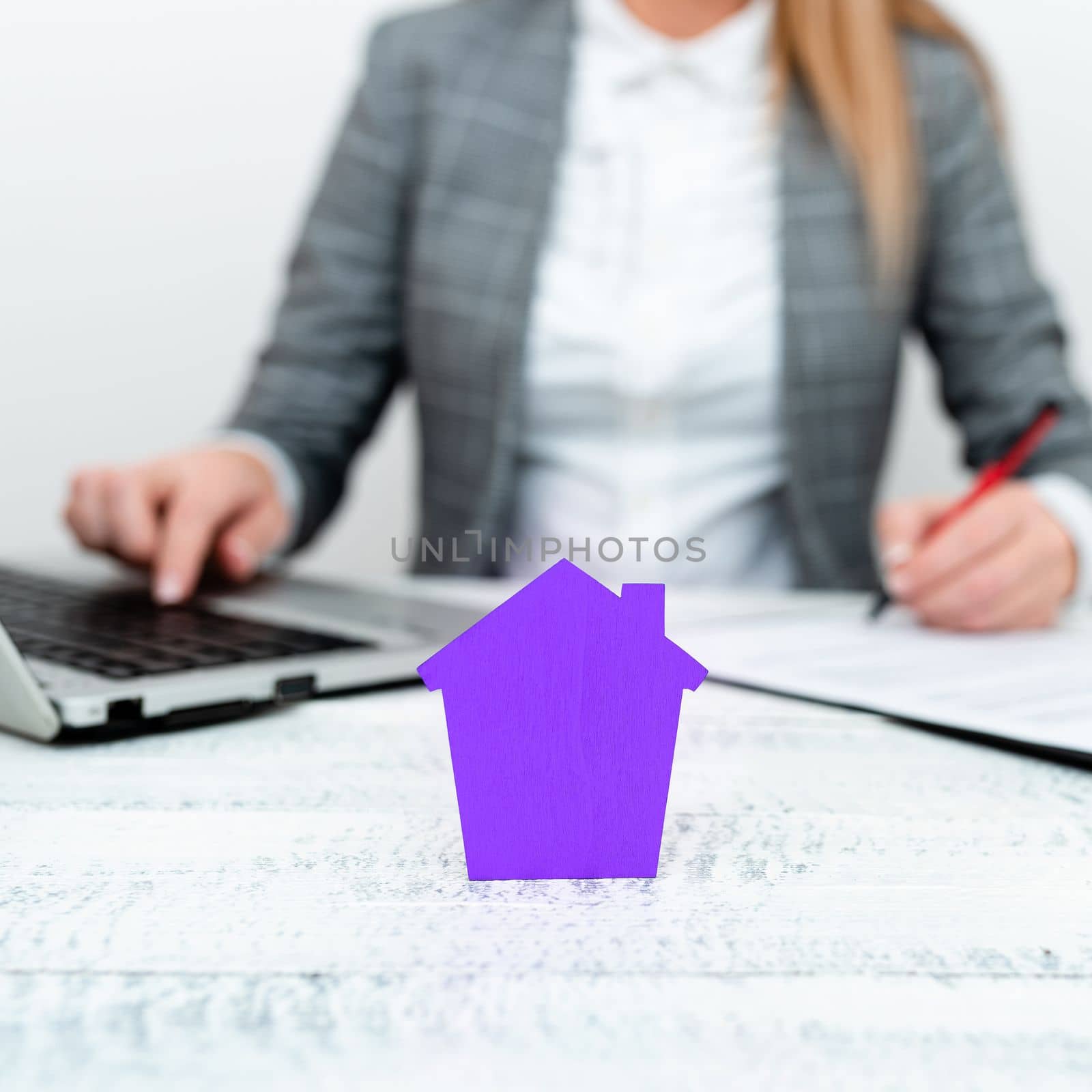 Businesswoman sitting at table writing in notebook and typing on laptop. olored paper house standing on desk. Display important information. Display the latest news. by nialowwa