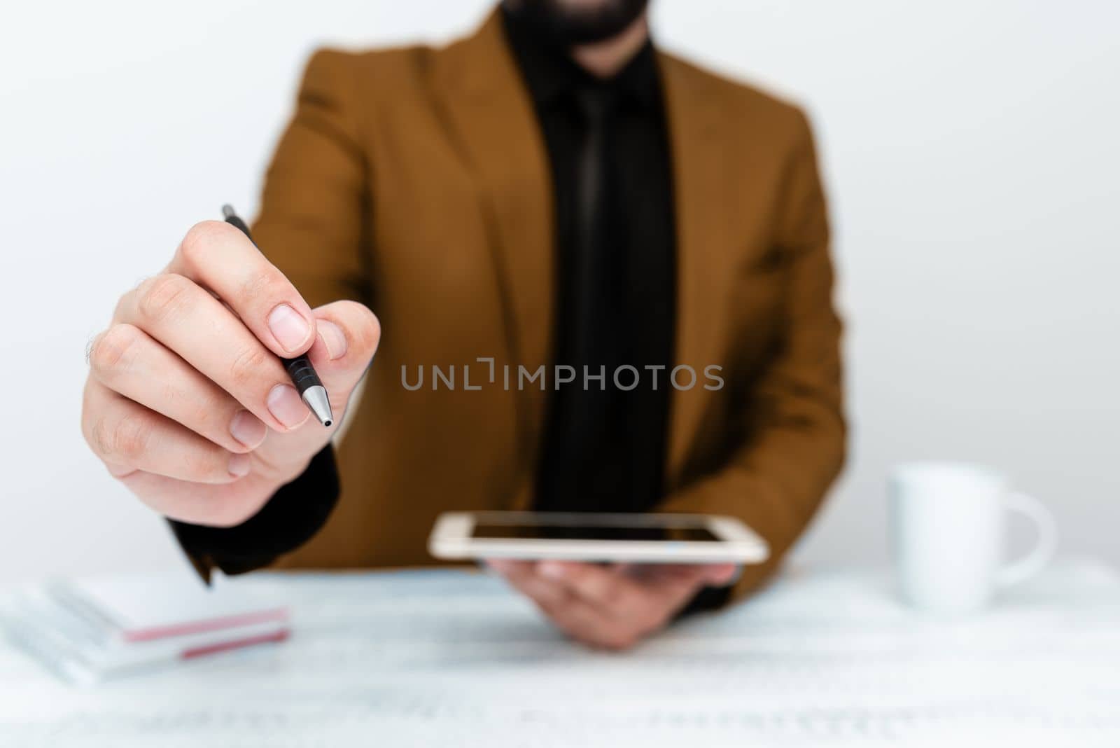 Man in Brown jacket sitting at white table And Pointing With Pen On Important Message. Holding mobile phone. Gentleman Showing Critical Announcement. by nialowwa