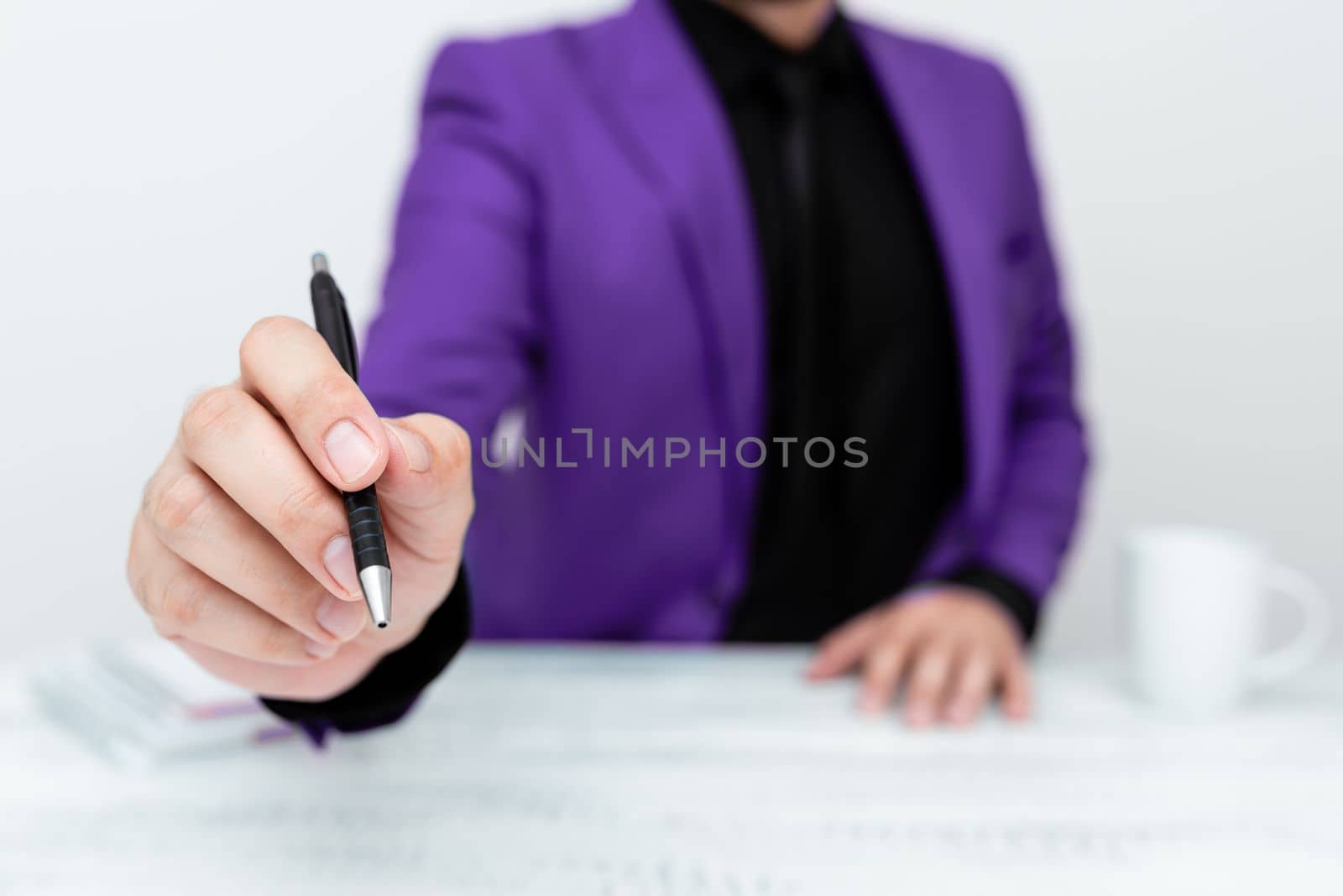 Male model in Purple suit sitting at white table And Pointing With Pen On Important Message. Gentleman Showing Critical Announcement. Coffee cup on deck. by nialowwa