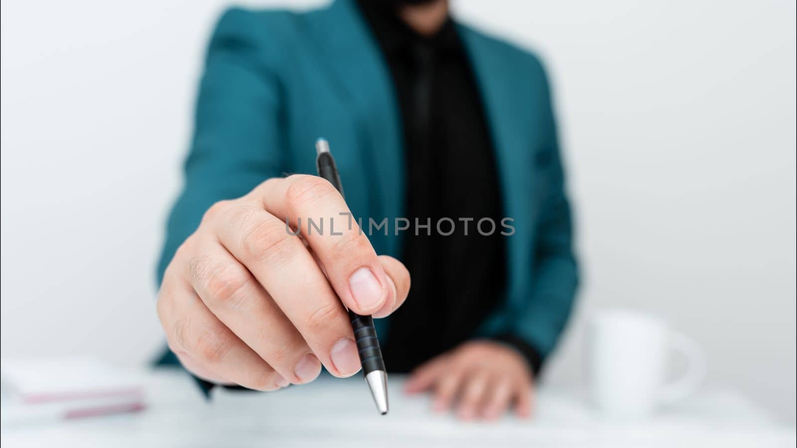 Male model in Blue suit sitting at white table And Pointing With Pen On Important Message. Gentleman Showing Critical Announcement. Coffee cup on deck. by nialowwa