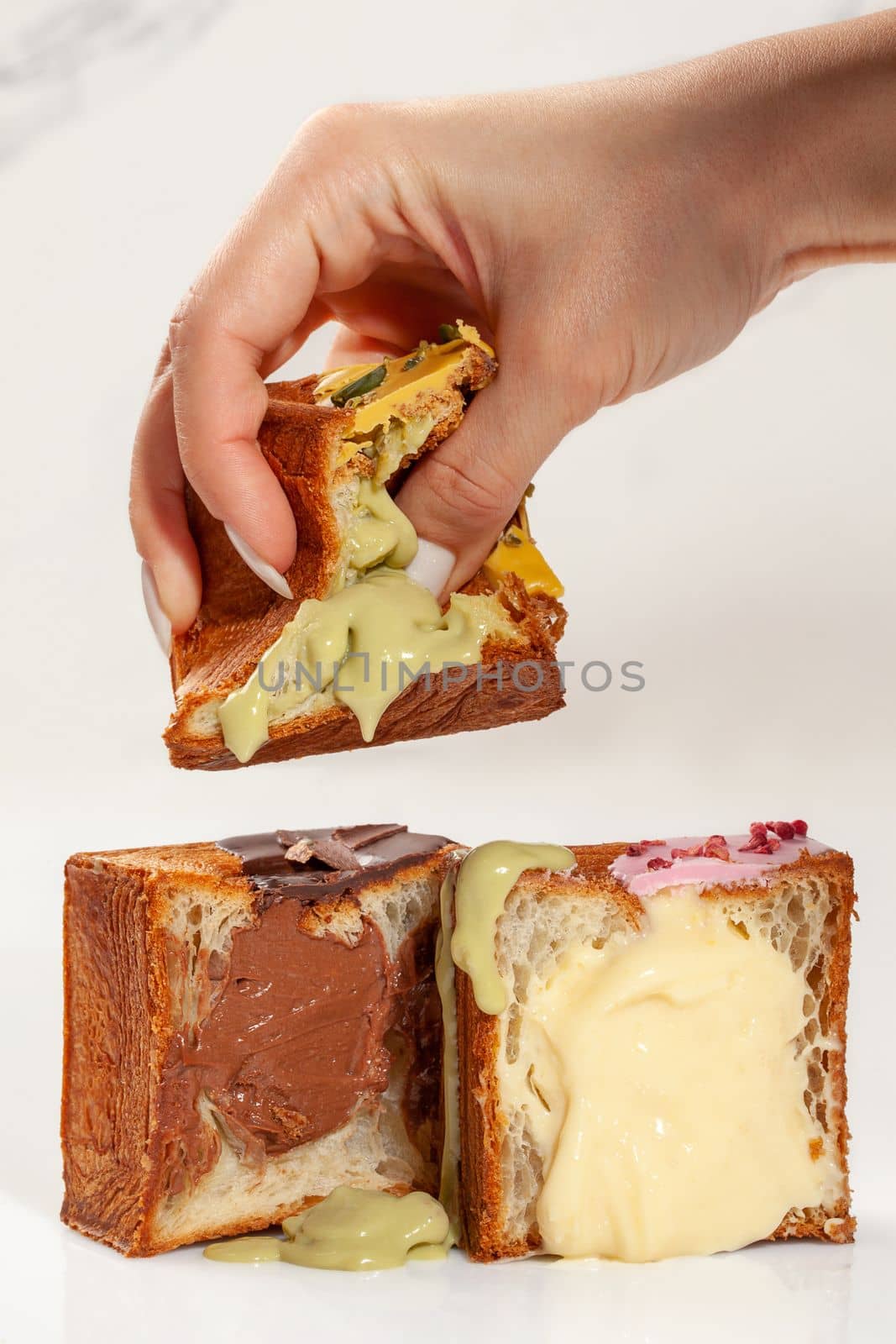 Woman squeezing with fingers soft airy freshly baked cubic croissant with pistachio custard while two more halves with various sweet fillings lying on table, cropped image. Sweet tooth concept