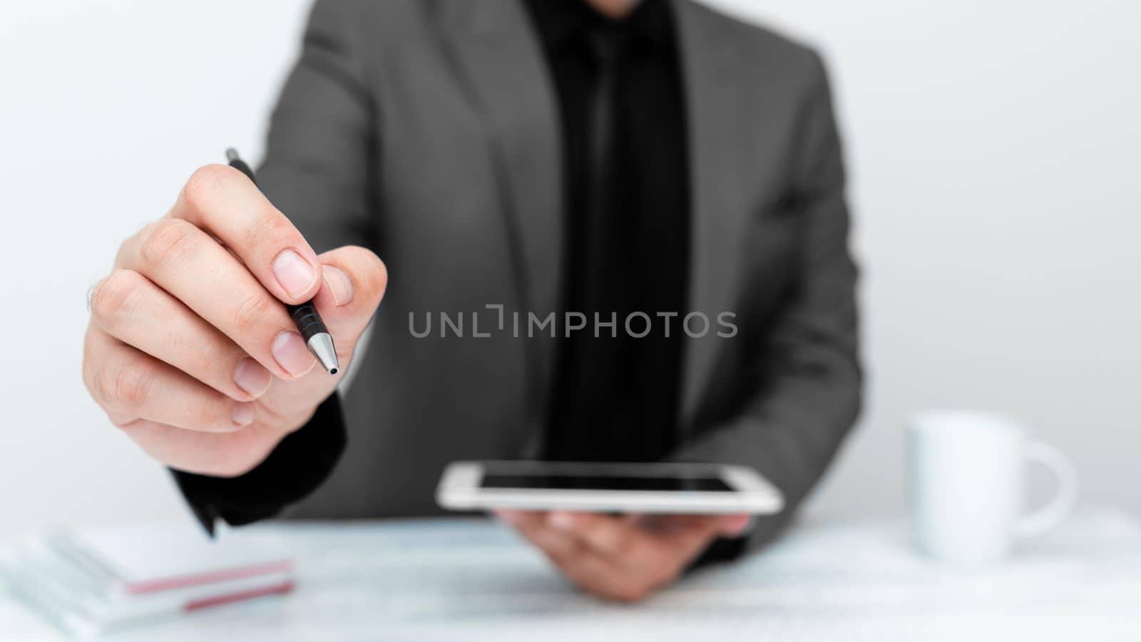 Man in Gray jacket sitting at white table And Pointing With Pen On Important Message. Holding mobile phone. Gentleman Showing Critical Announcement. by nialowwa