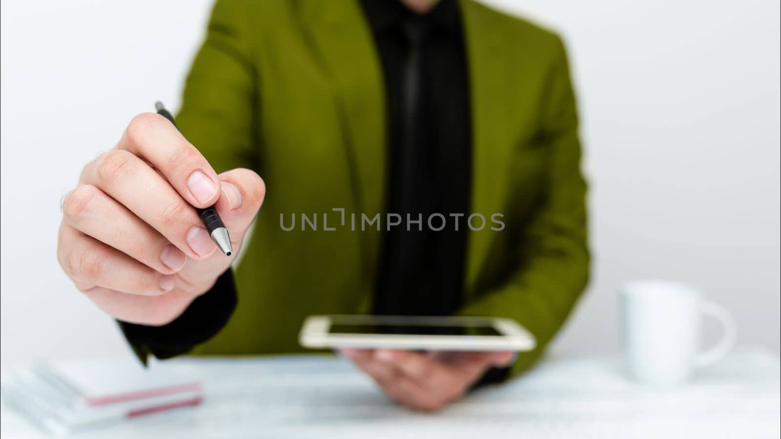 Man in Green jacket sitting at white table And Pointing With Pen On Important Message. Holding mobile phone. Gentleman Showing Critical Announcement. by nialowwa