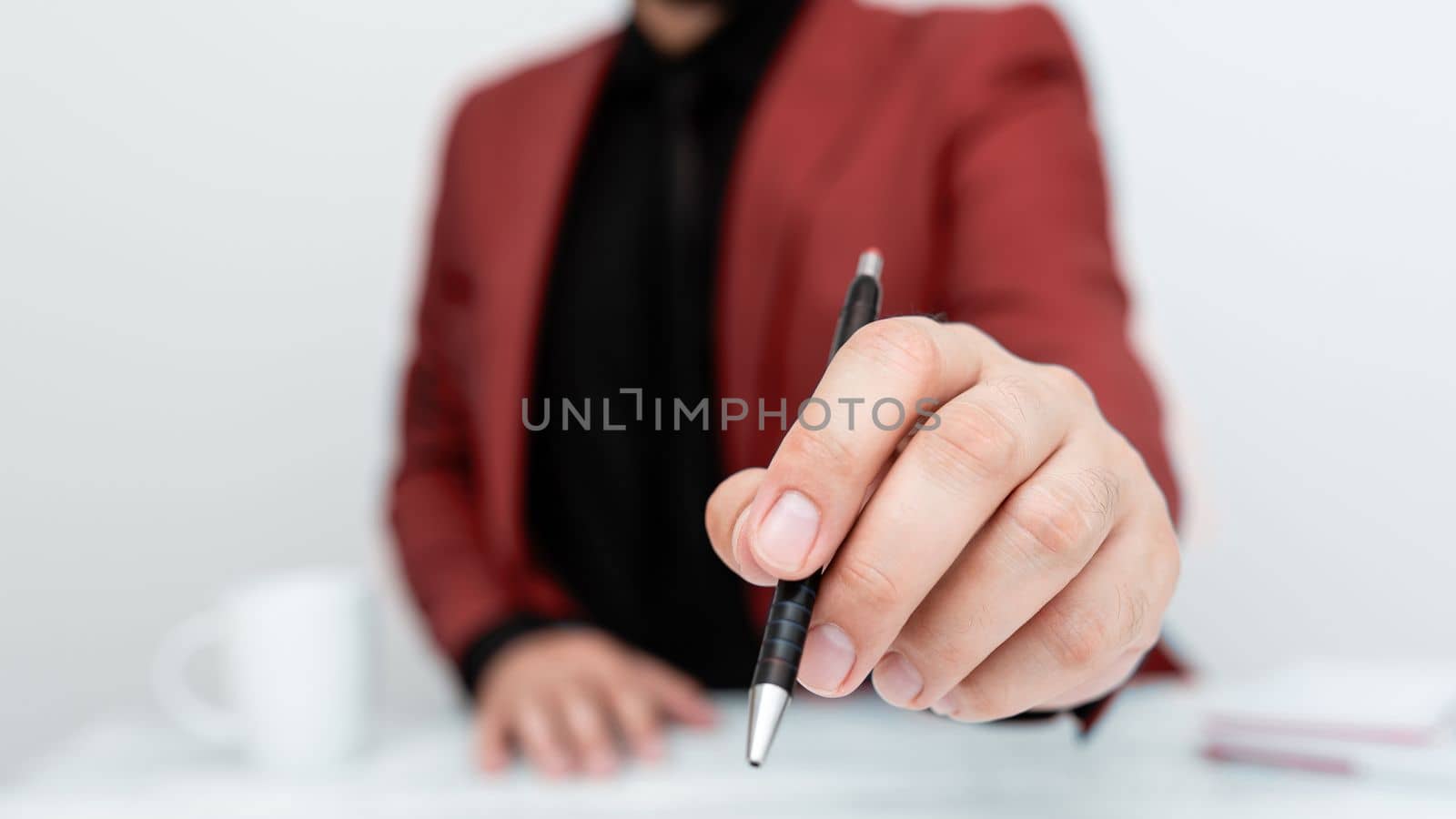 Male model in Red suit sitting at white table And Pointing With Pen On Important Message. Gentleman Showing Critical Announcement. Coffee cup on deck. by nialowwa