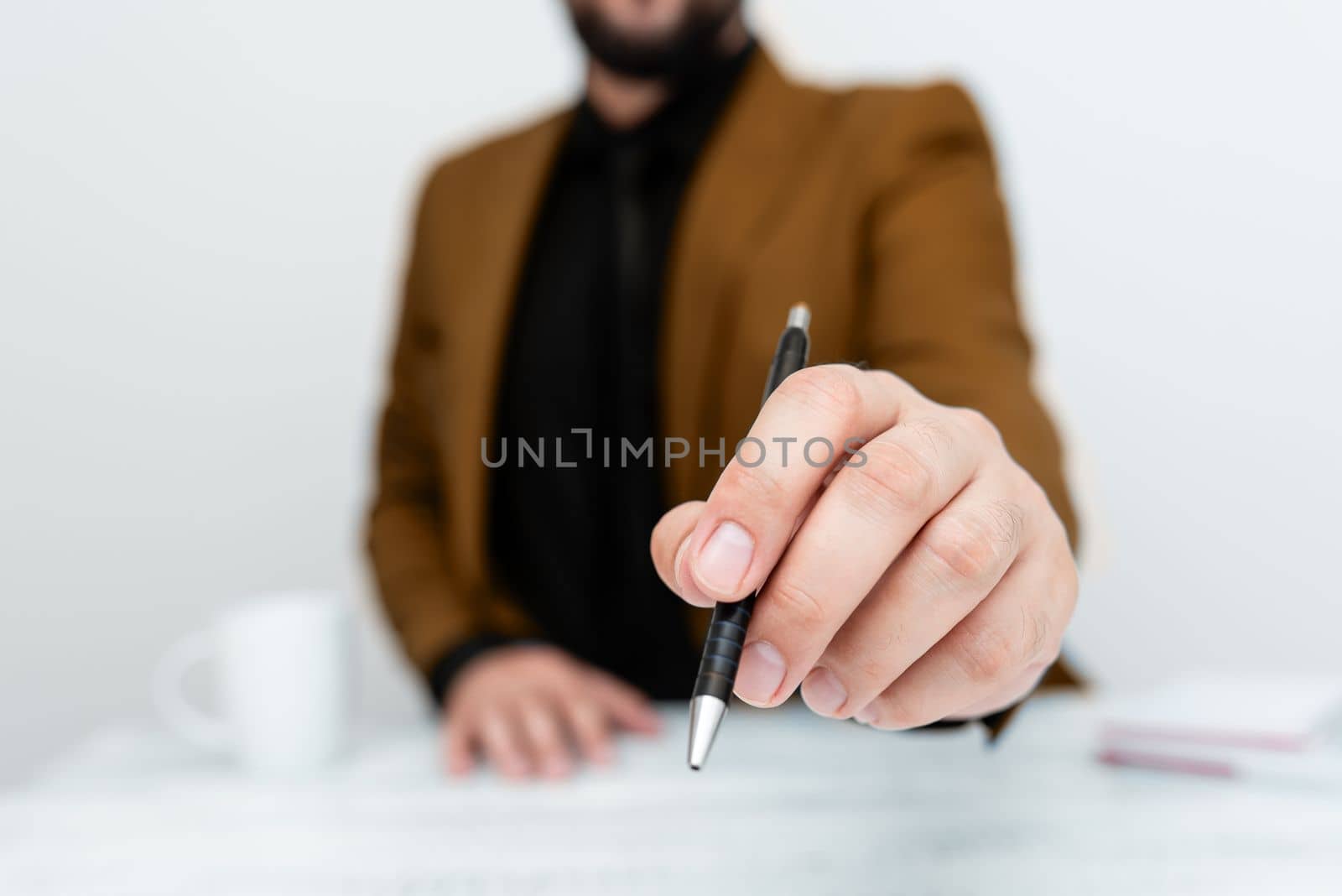 Male model in Brown suit sitting at white table And Pointing With Pen On Important Message. Gentleman Showing Critical Announcement. Coffee cup on deck. by nialowwa