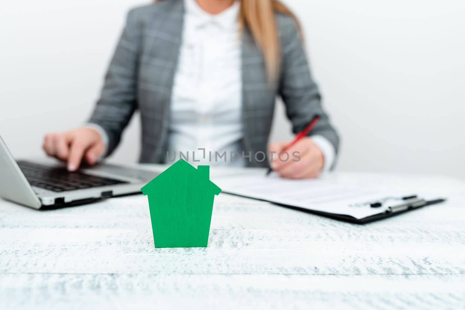 Businesswoman sitting at table writing in notebook and typing on laptop. olored paper house standing on desk. Display important information. Display the latest news. by nialowwa