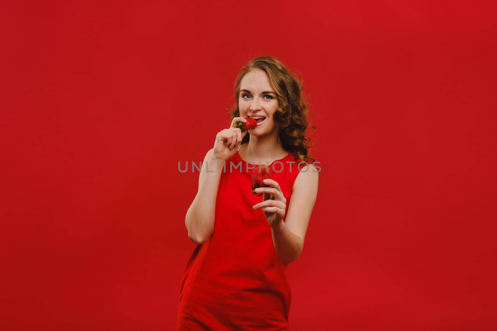 A beautiful girl in a red dress on a red background holds a strawberry in her hands and smiles by Lobachad