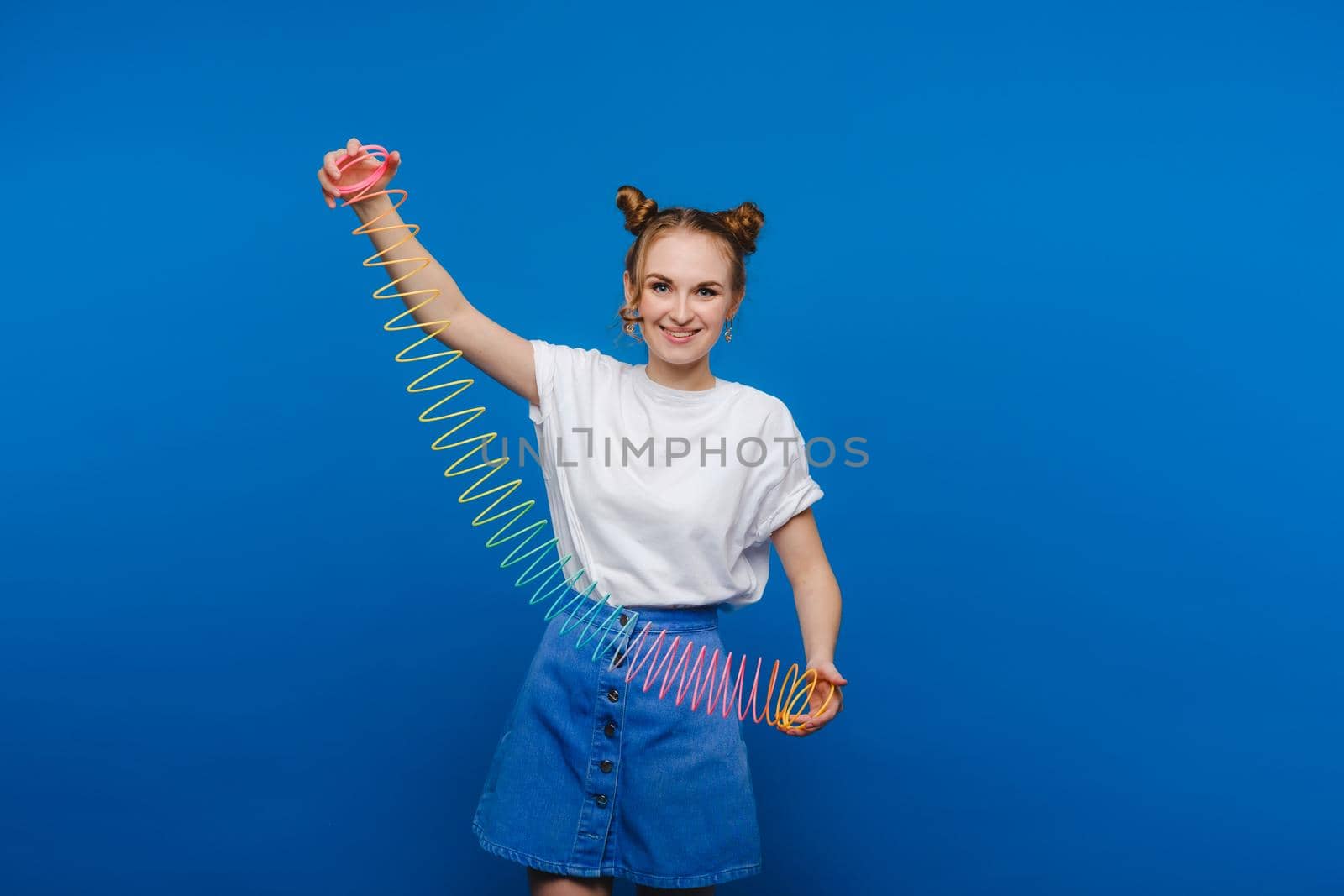 Beautiful young girl playing with a rainbow slinky, a toy of her childhood on a blue background by Lobachad