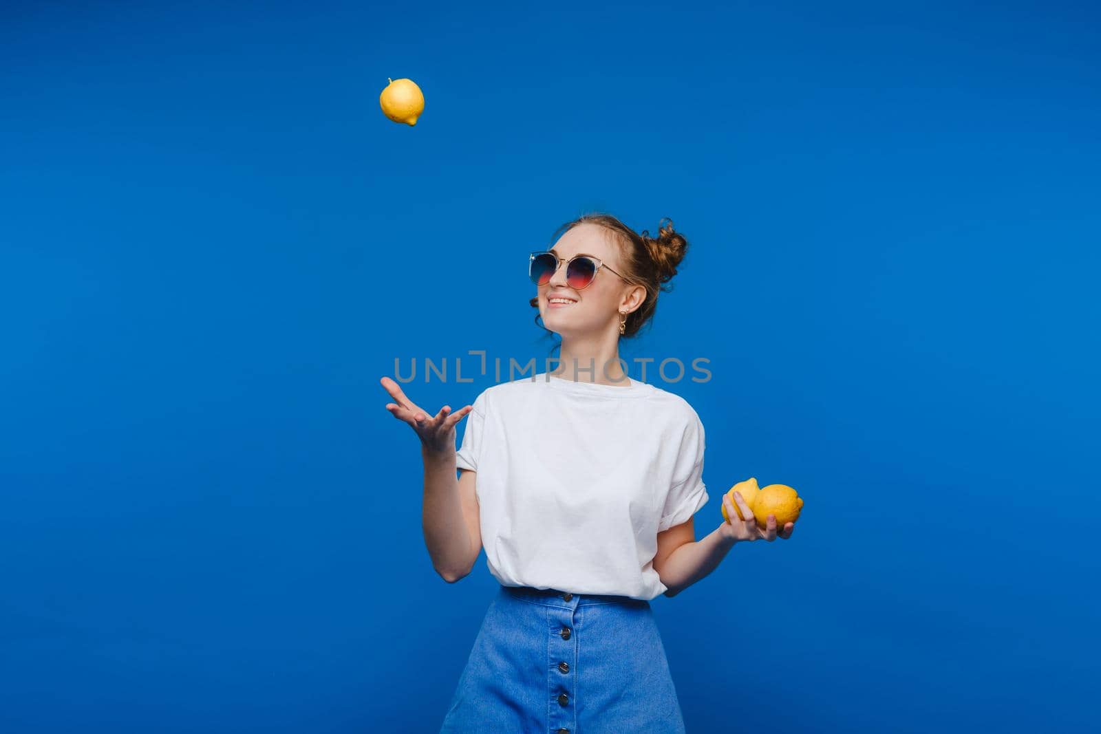 a young beautiful girl standing on a blue background holding lemons in her hand. Smiles by Lobachad