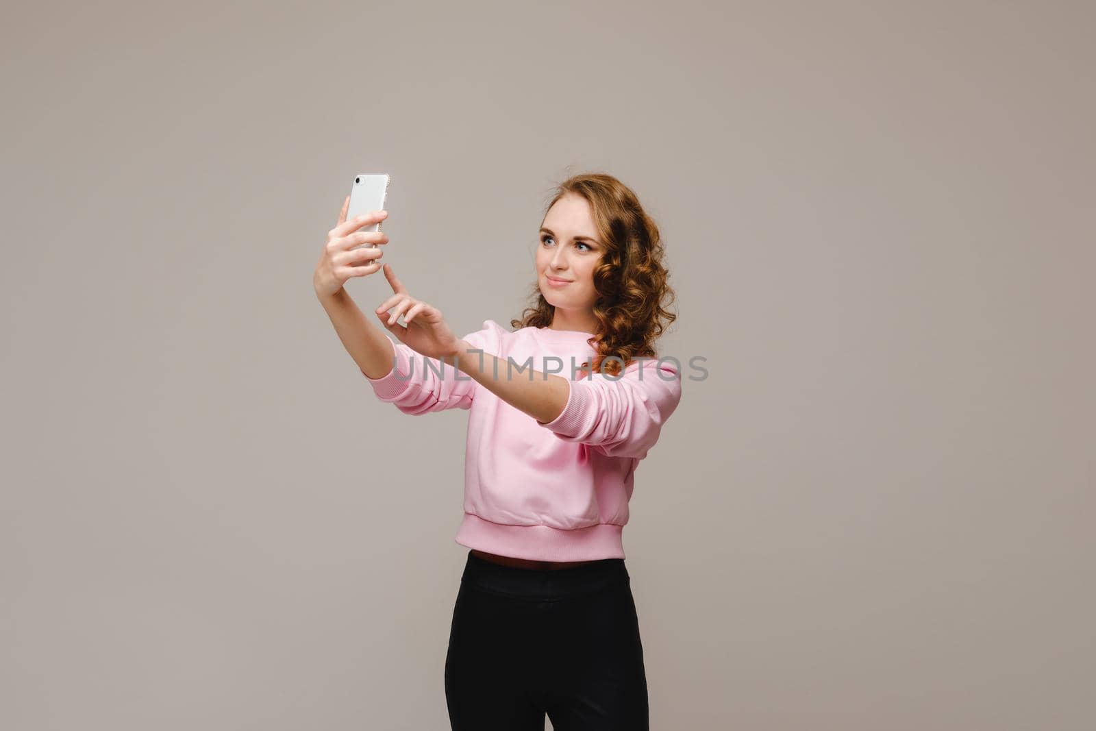 A smiling happy girl in a pink blouse takes a selfie on a smartphone on a gray background. by Lobachad