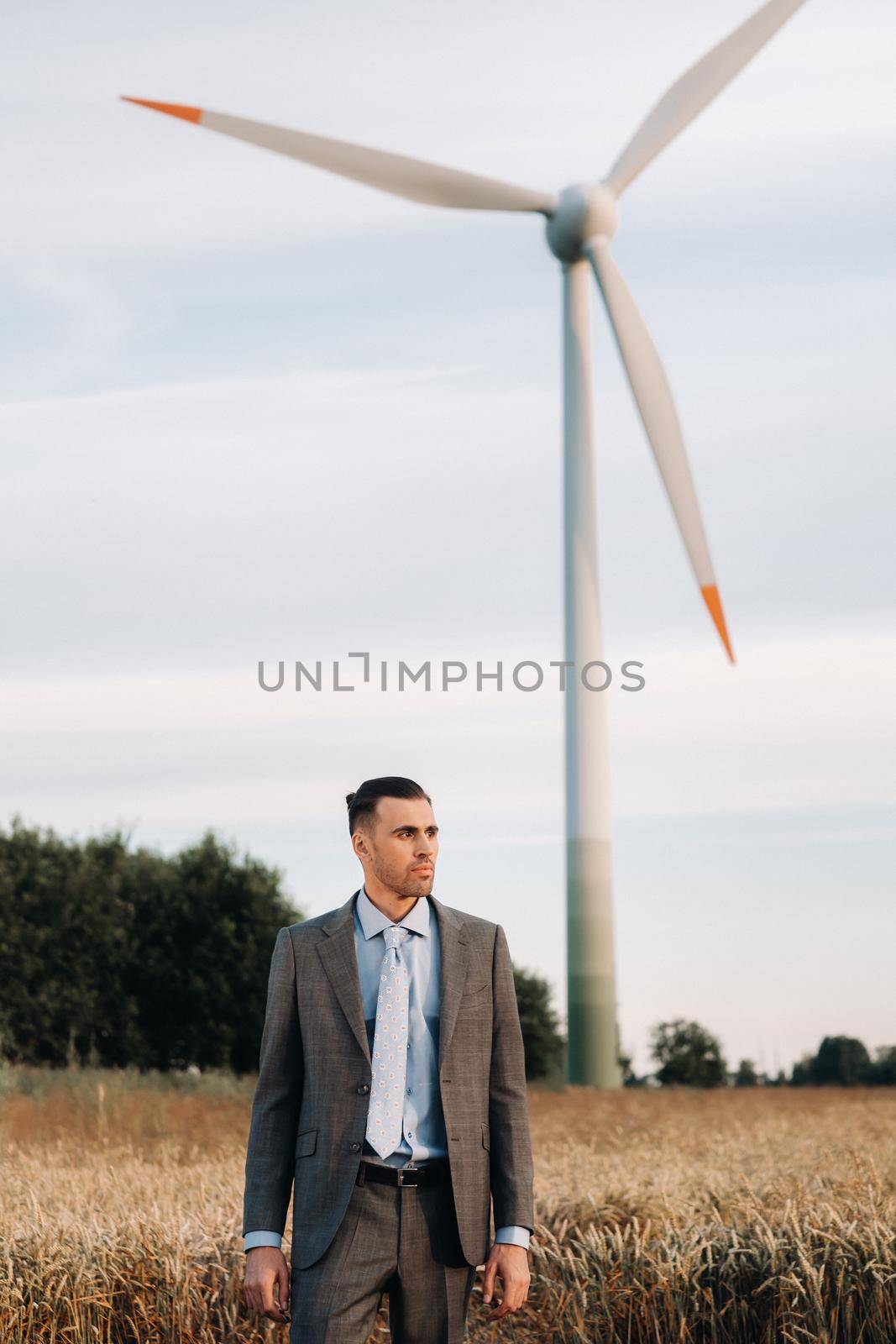 Portrait of a businessman in a gray suit on a wheat field against the background of a windmill and the evening sky.