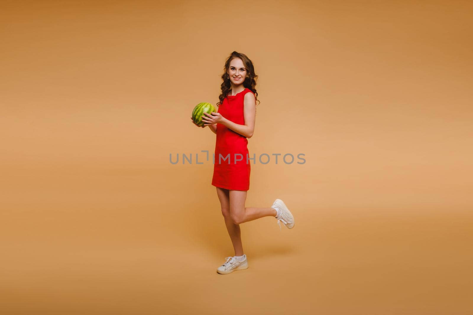 A beautiful girl in glasses and a red dress holds a watermelon in her hands.