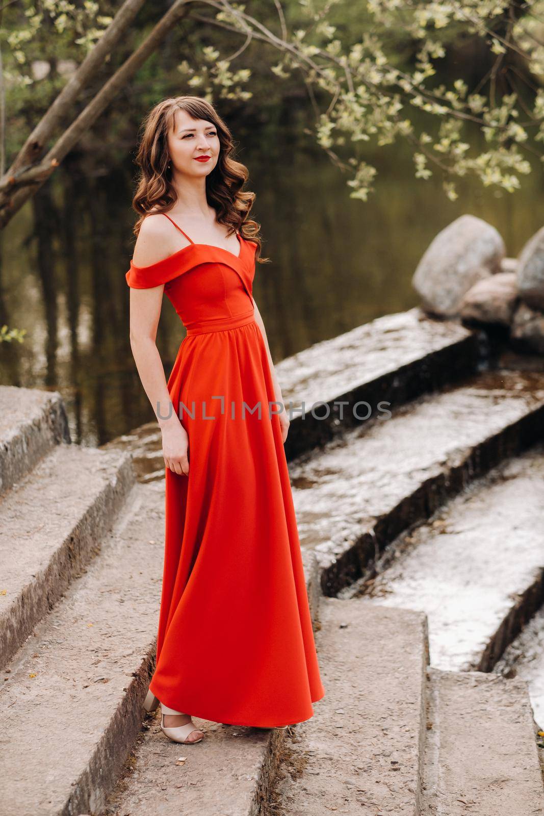 a young beautiful girl with long brown hair , in a long red dress comes down the stairs near the lake,
