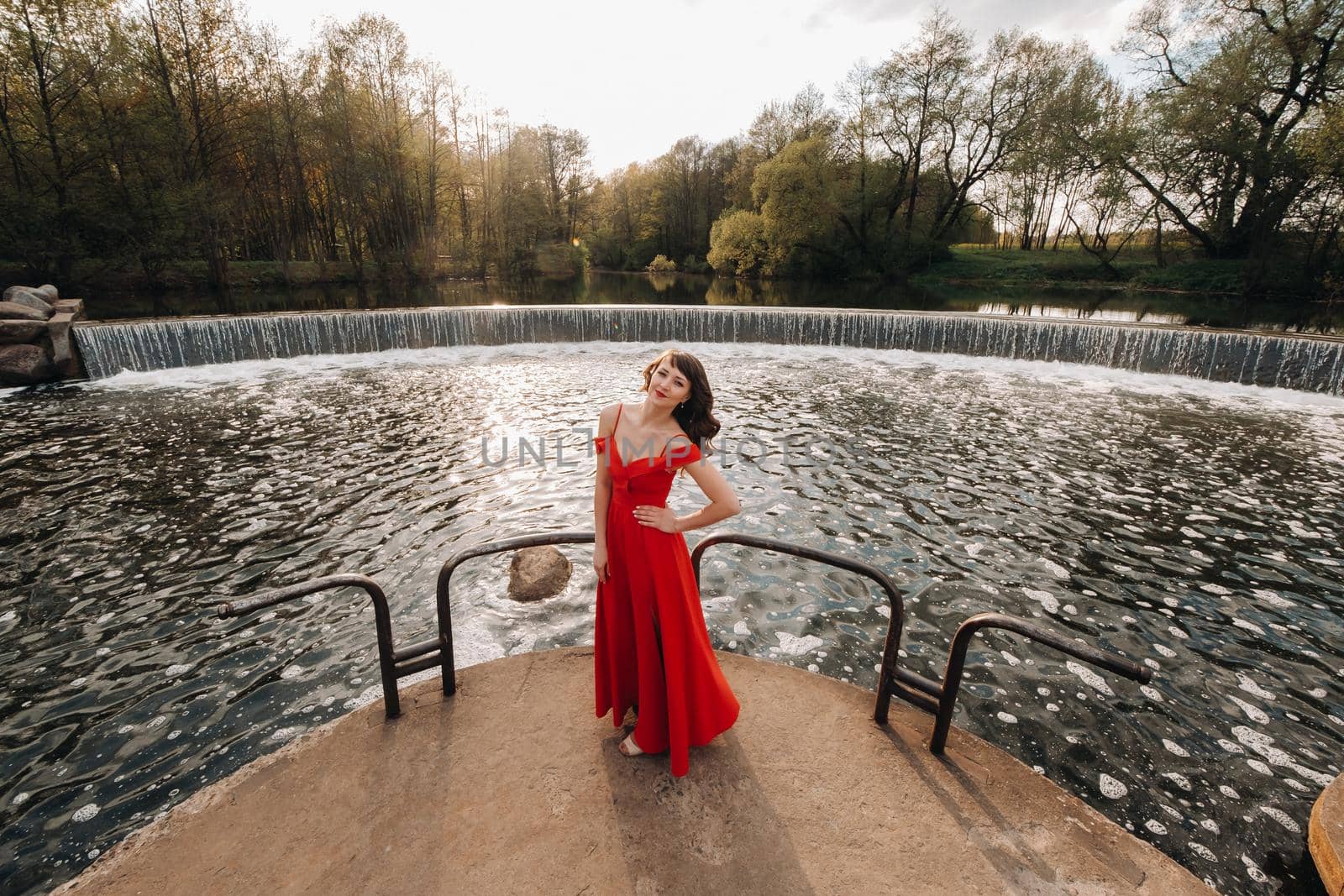 girl in a long red dress near the lake in the Park at sunset. by Lobachad