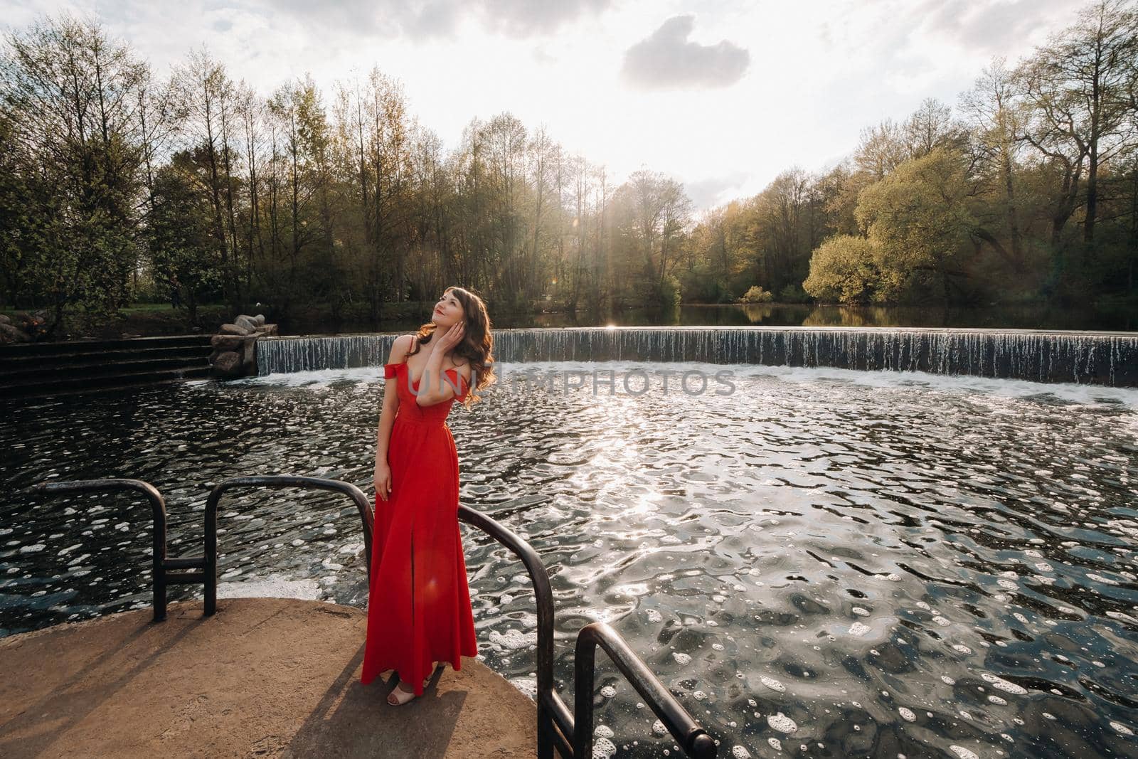 girl in a long red dress near the lake in the Park at sunset. by Lobachad