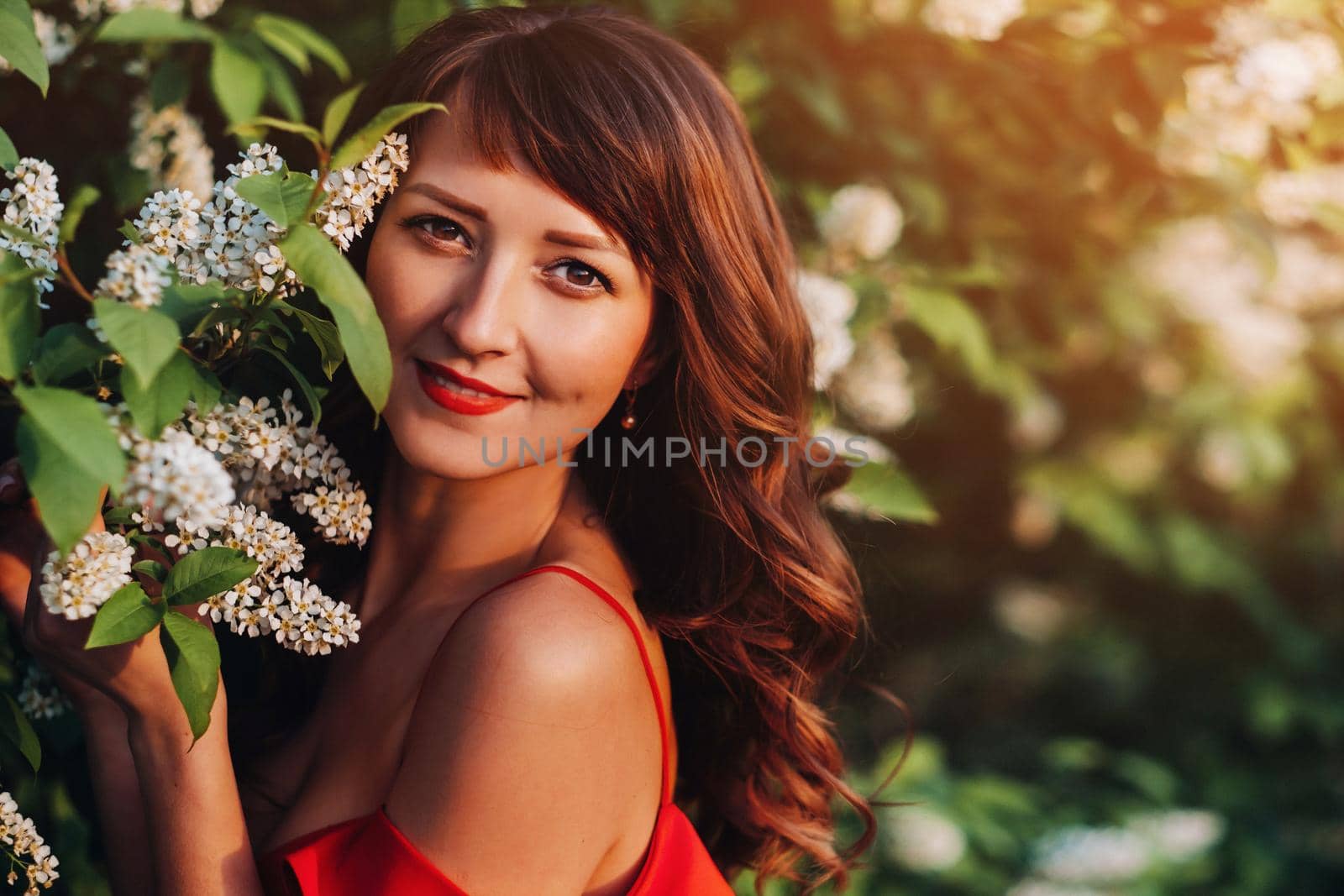 a girl in a red dress with red lips stands next to a large white flowering tree At sunset by Lobachad