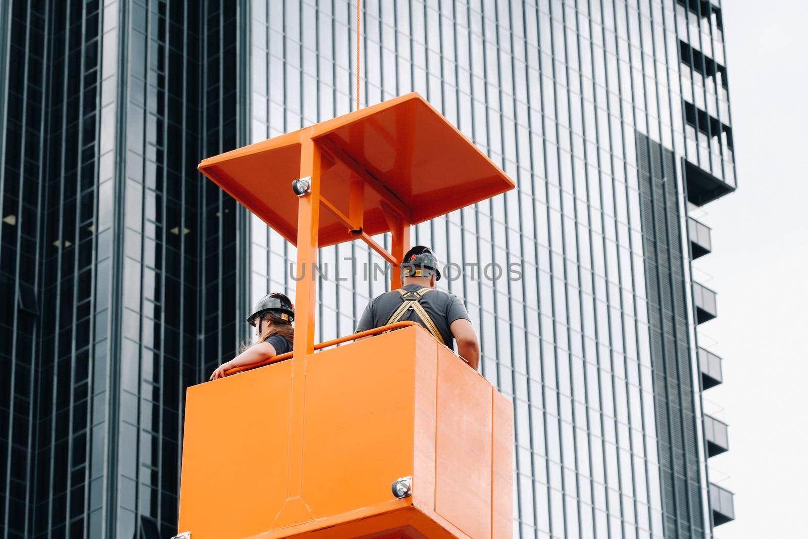 Workers in a construction cradle climb on a crane to a large glass building.The crane lifts the workers in the car seat.Construction.