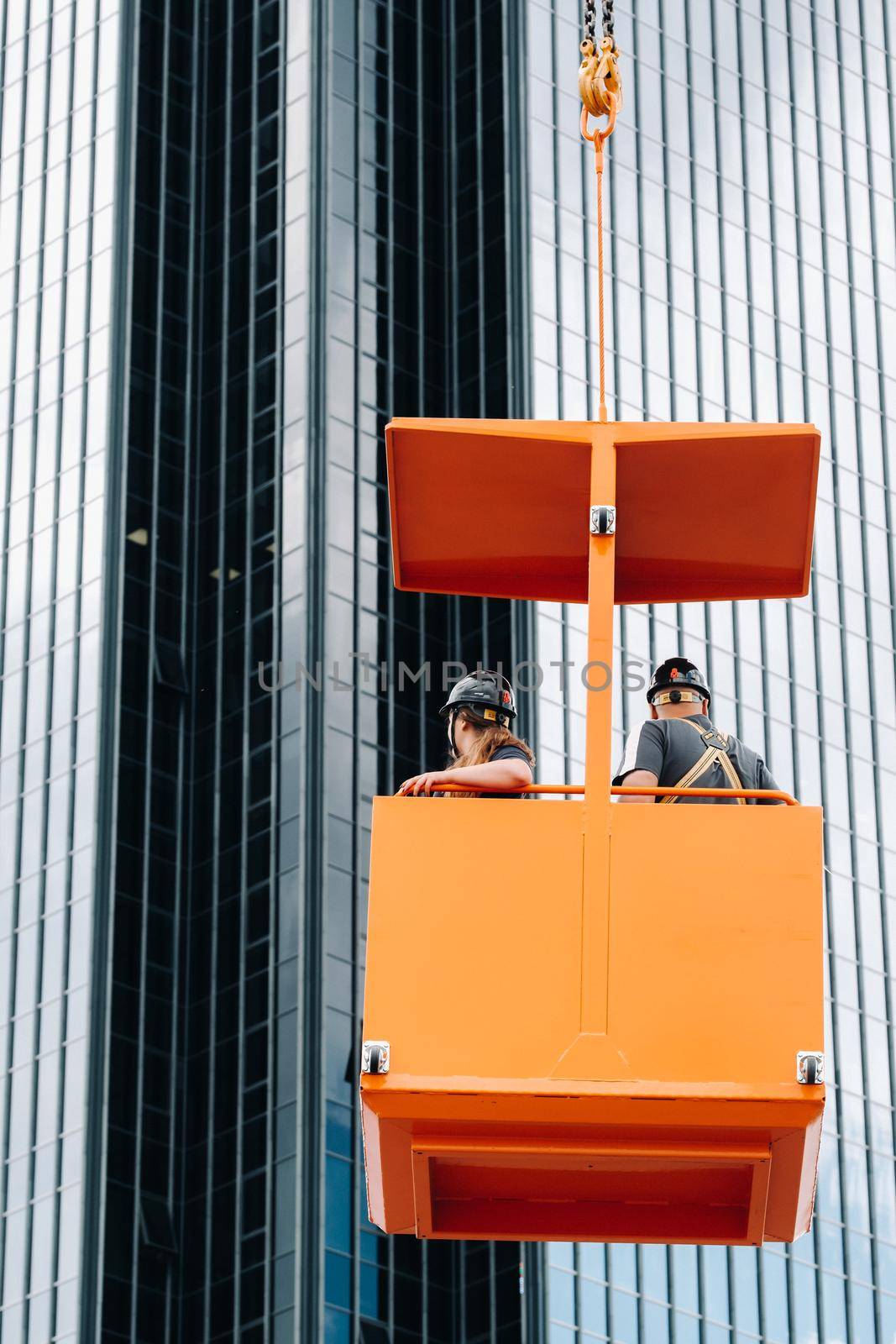 Workers in a construction cradle climb on a crane to a large glass building.The crane lifts the workers in the car seat.Construction.