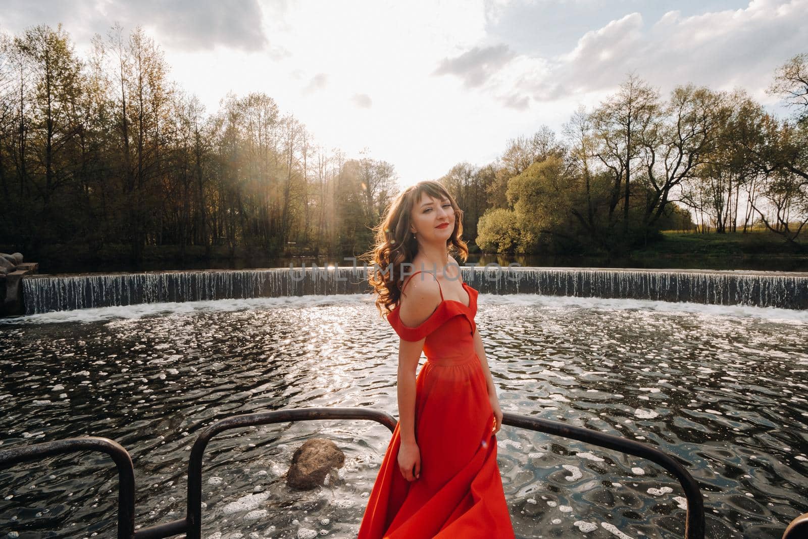 girl in a long red dress near the lake in the Park at sunset. by Lobachad