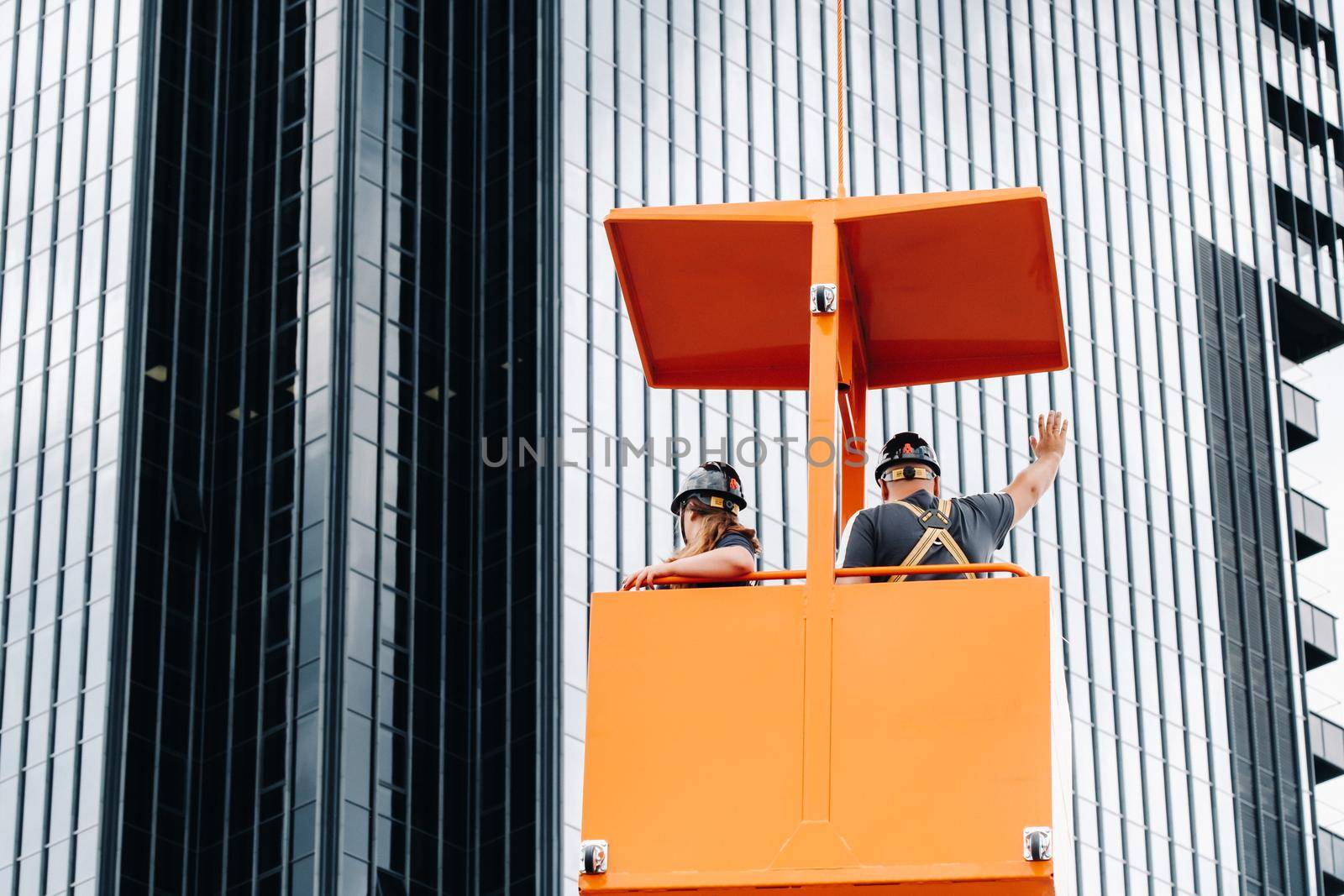Workers in a construction cradle climb on a crane to a large glass building.The crane lifts the workers in the car seat.Construction by Lobachad