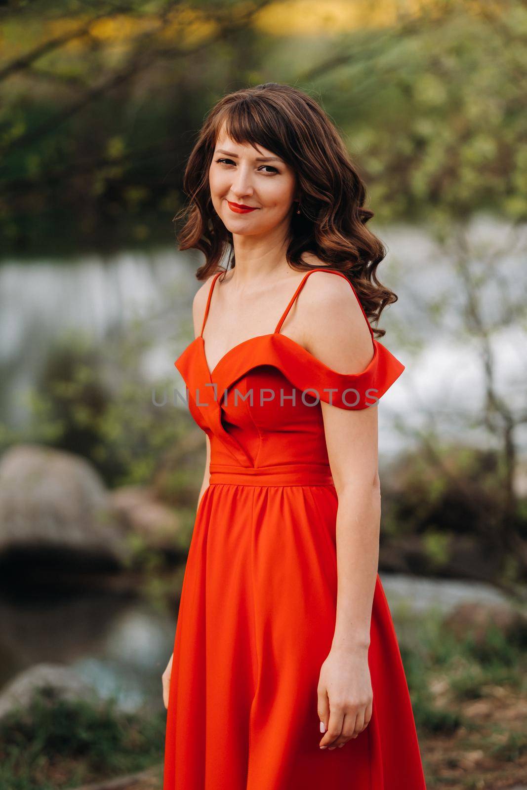 girl in a long red dress near the lake in the Park at sunset. by Lobachad