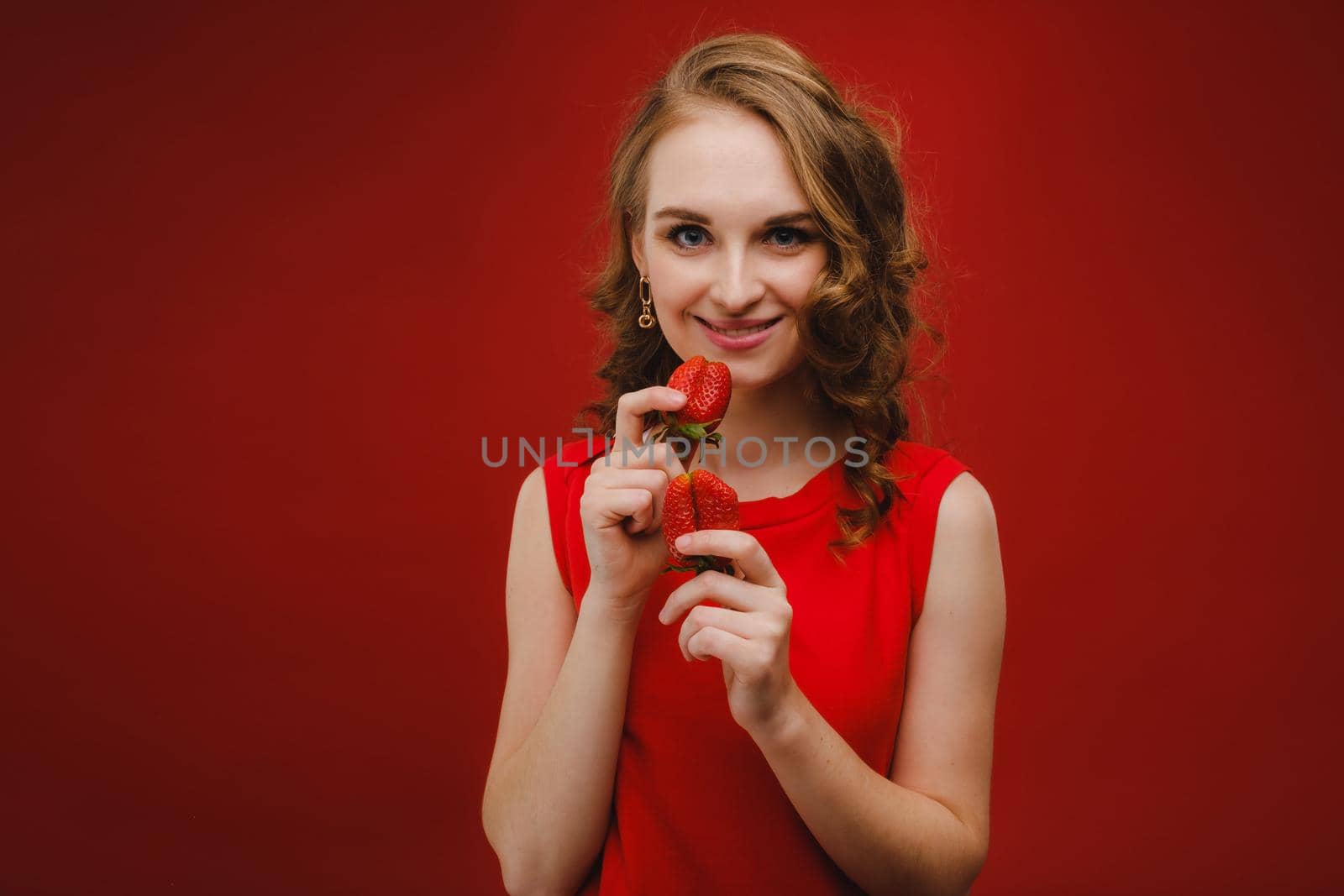 A beautiful girl in a red dress on a red background holds a strawberry in her hands and smiles.