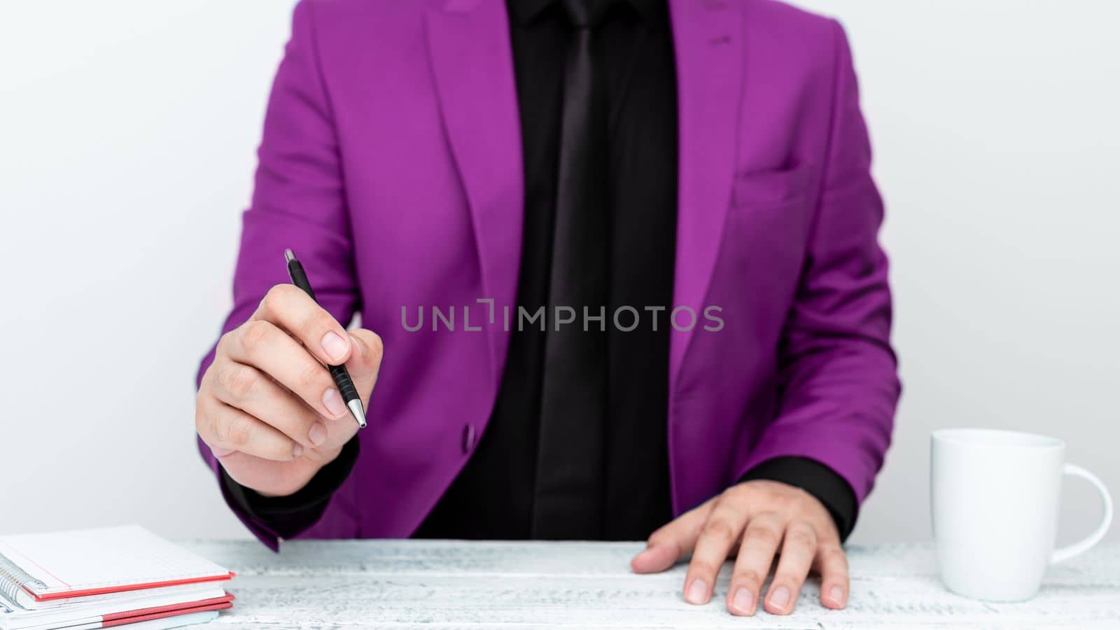 Male model in Pink suit sitting at white table And Pointing With Pen On Important Message. Gentleman Showing Critical Announcement. Coffee cup on deck. by nialowwa