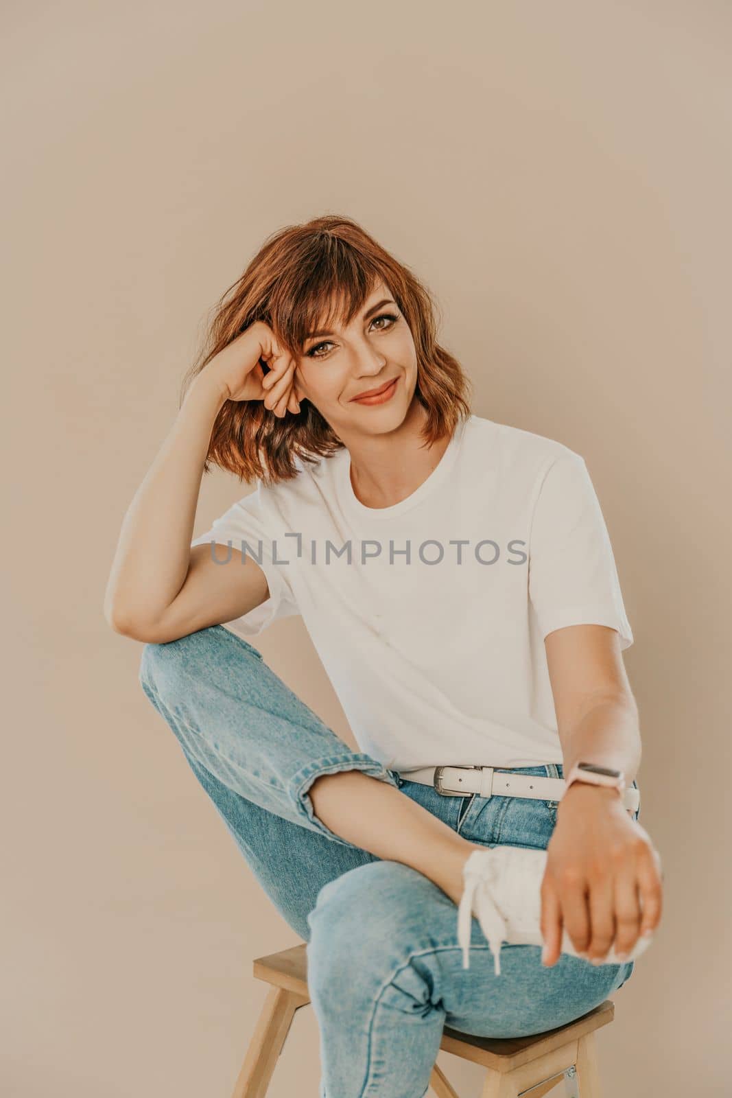 Portrait of brunette white t-shirt. A woman sits on a chair and looks at the camera, business portrait on a beige background, dressed in a white t-shirt and jeans. by Matiunina