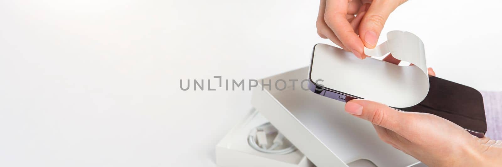 Unpacking a new phone. Remove the protective white film from the new phone. Close-up of a woman's hands removing a protective film from a phone in a purple case. Copy space on white background. by SERSOL